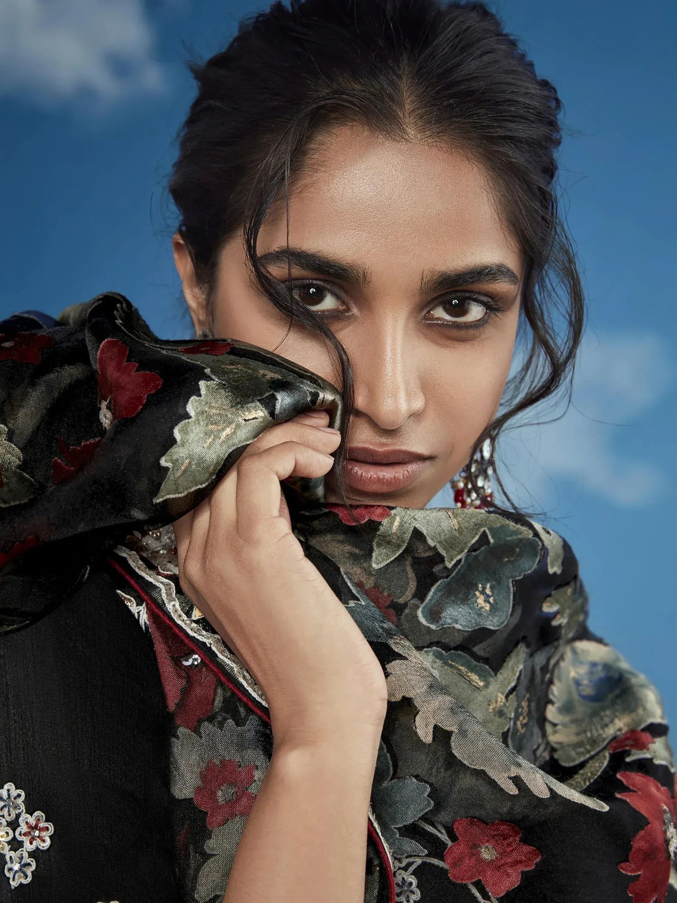 Woman in black floral ethnic wear with red and gold embroidery, holding dupatta against blue sky