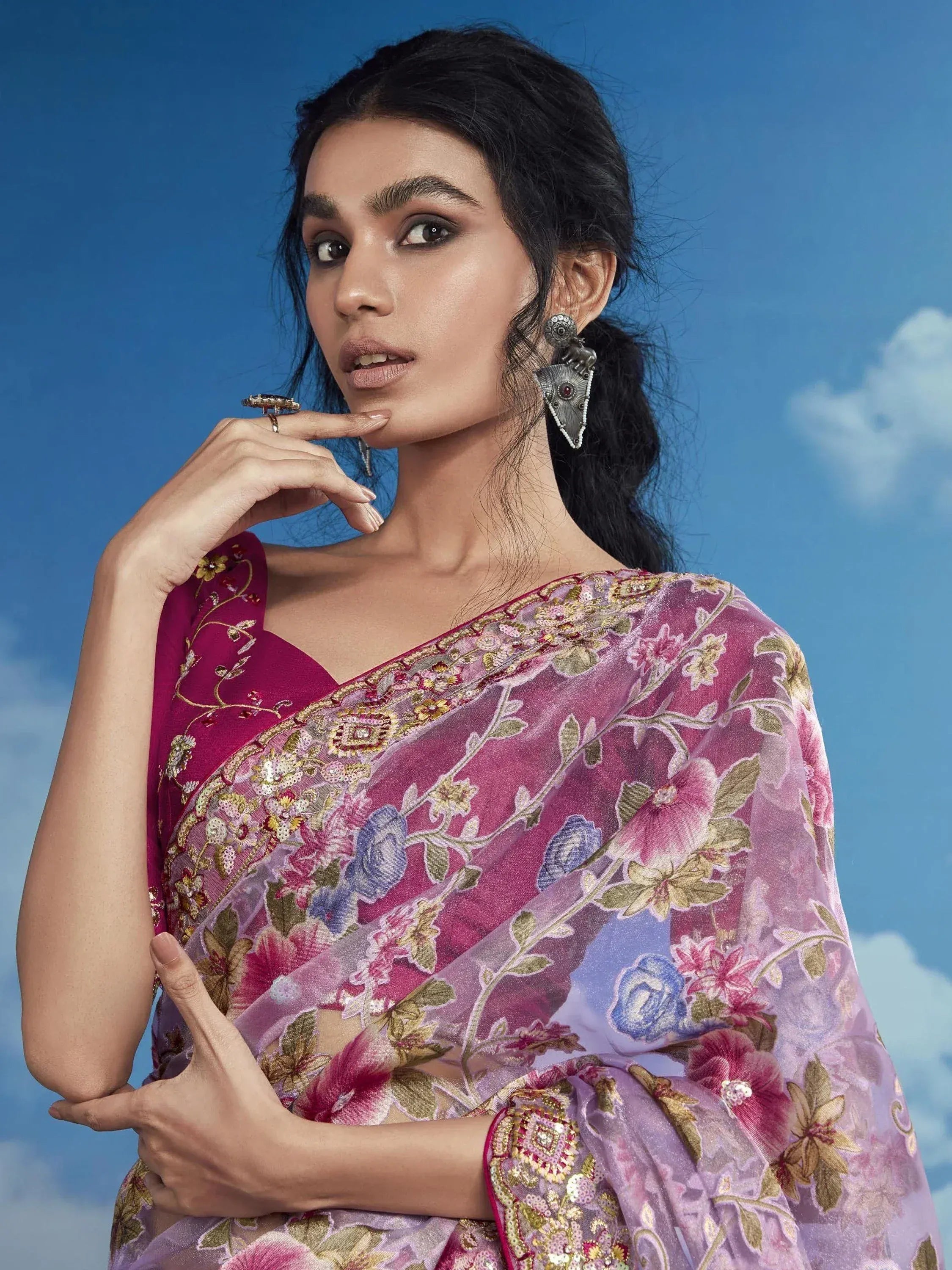 Woman in floral embroidered pink saree with gold detailing, styled with statement earrings against blue sky