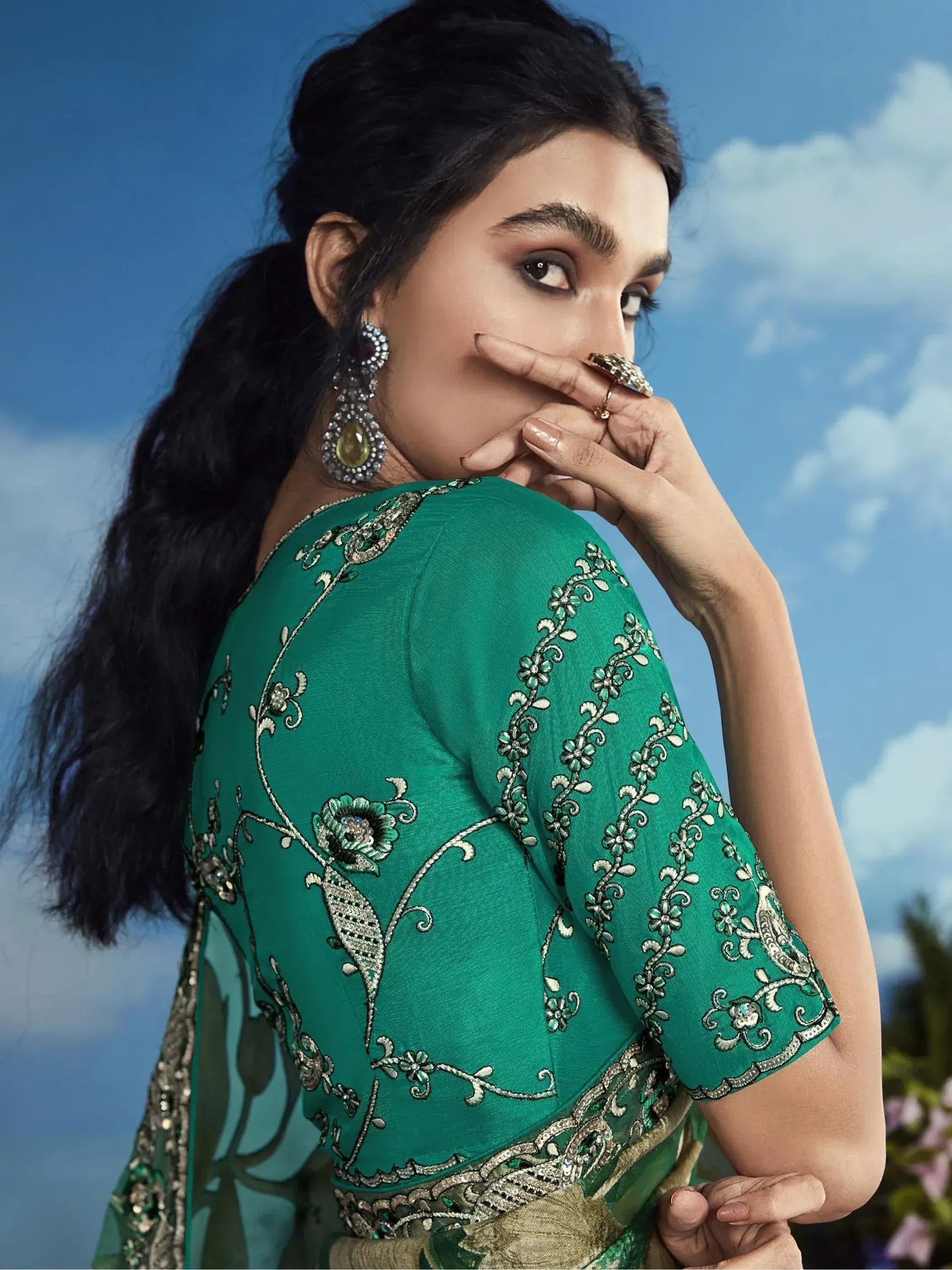 Woman in green embroidered Indian ethnic wear with floral design, statement earrings, and sky background