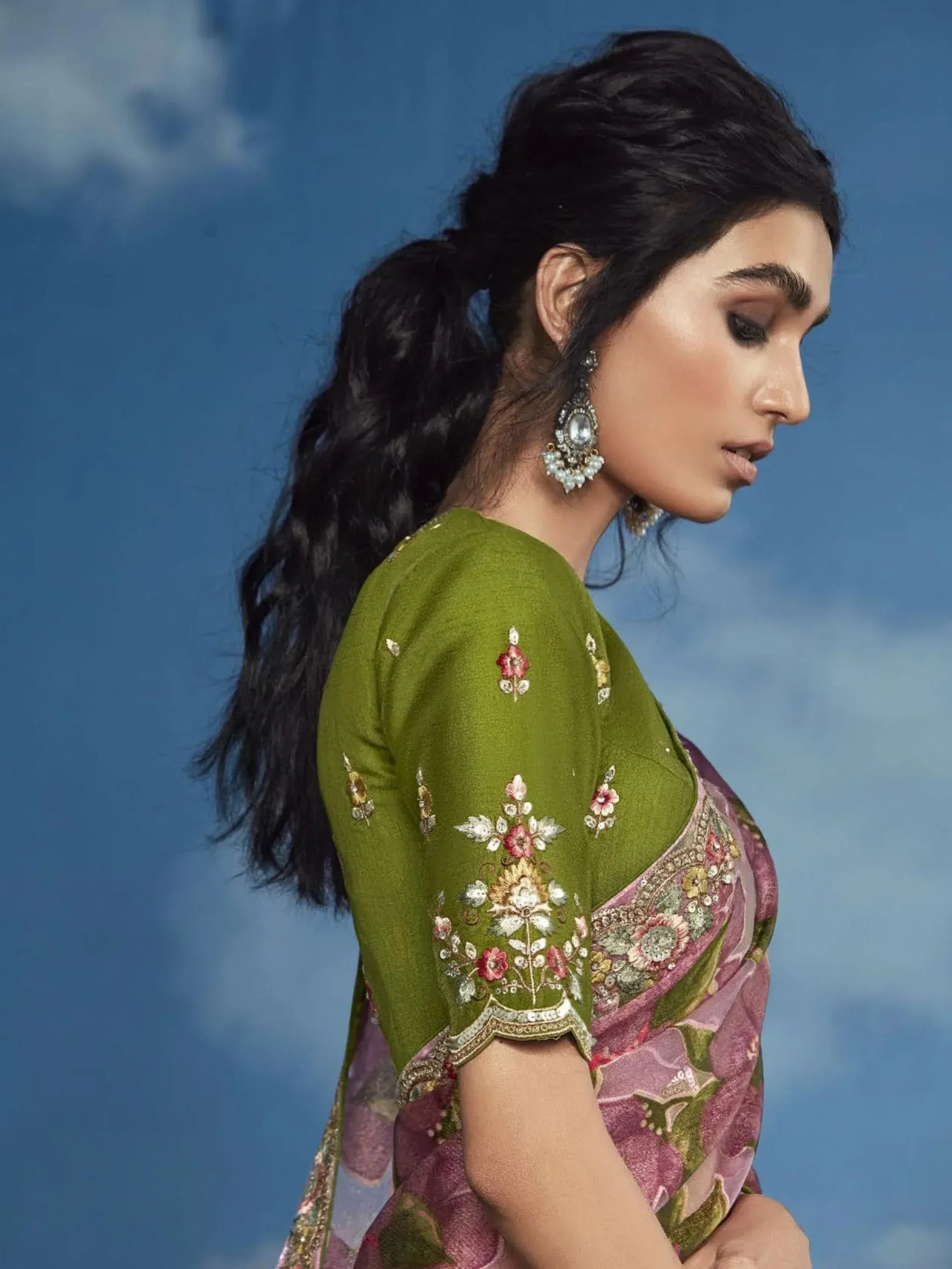 Woman in green embroidered saree blouse with floral design, paired with pink floral saree, against blue sky