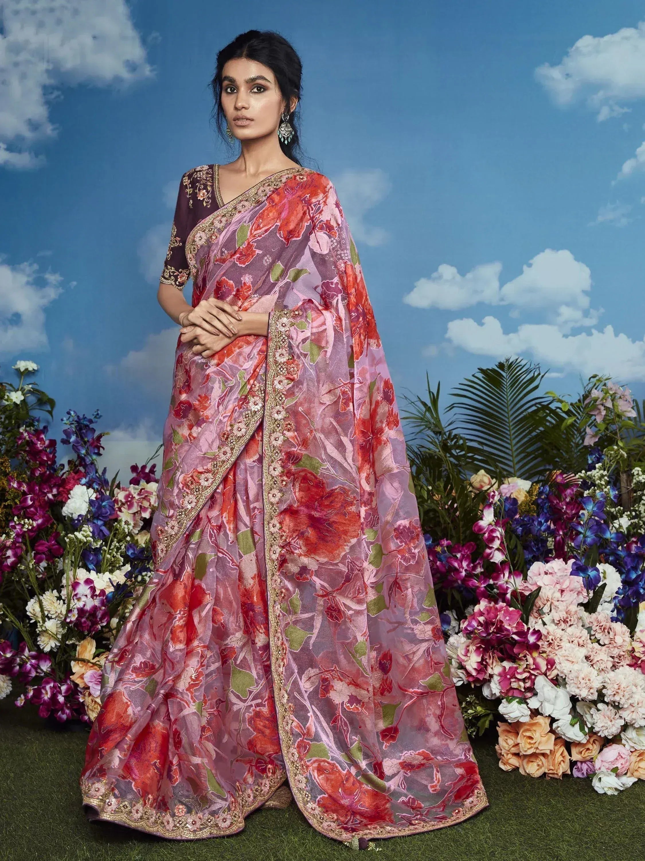 Woman in floral print Indian saree with gold embroidery, standing amidst colorful flowers under blue sky