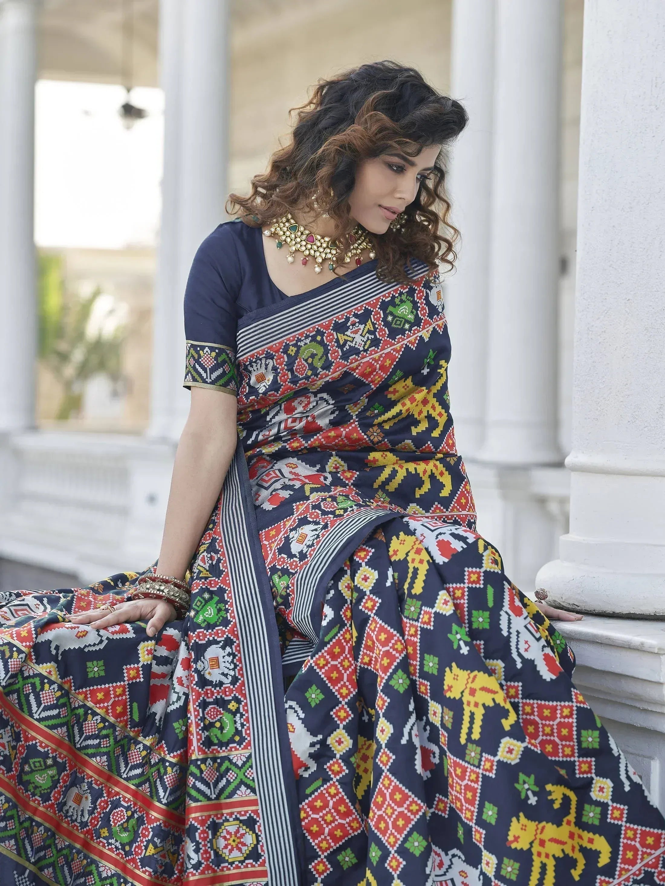 Woman in navy blue ethnic saree with colorful geometric and animal print, paired with gold jewelry, sitting near white pillars.