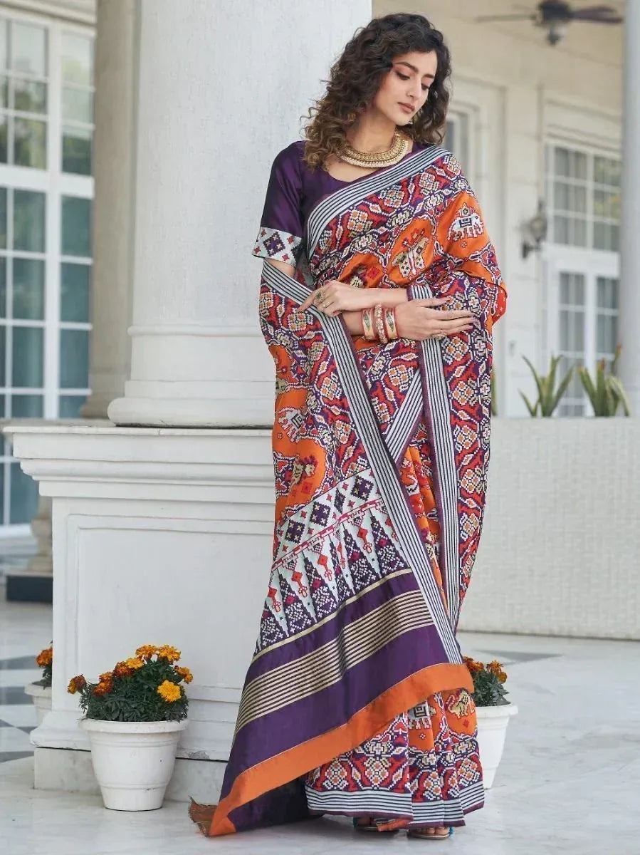 Woman in vibrant orange and purple printed saree with geometric patterns, styled with gold jewelry for ethnic occasions