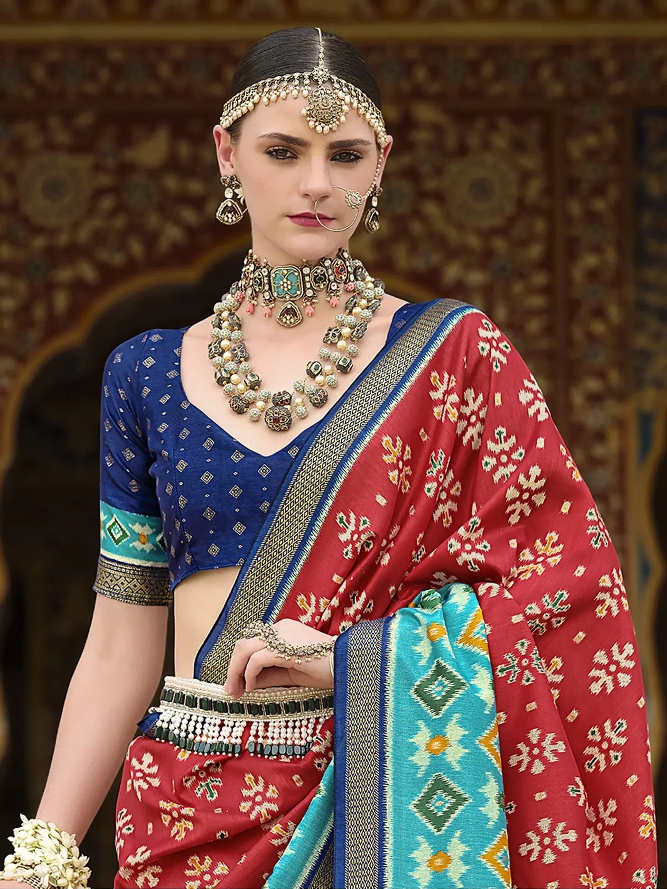 Red and blue ethnic saree with intricate patterns, paired with traditional jewelry and a matching blouse.