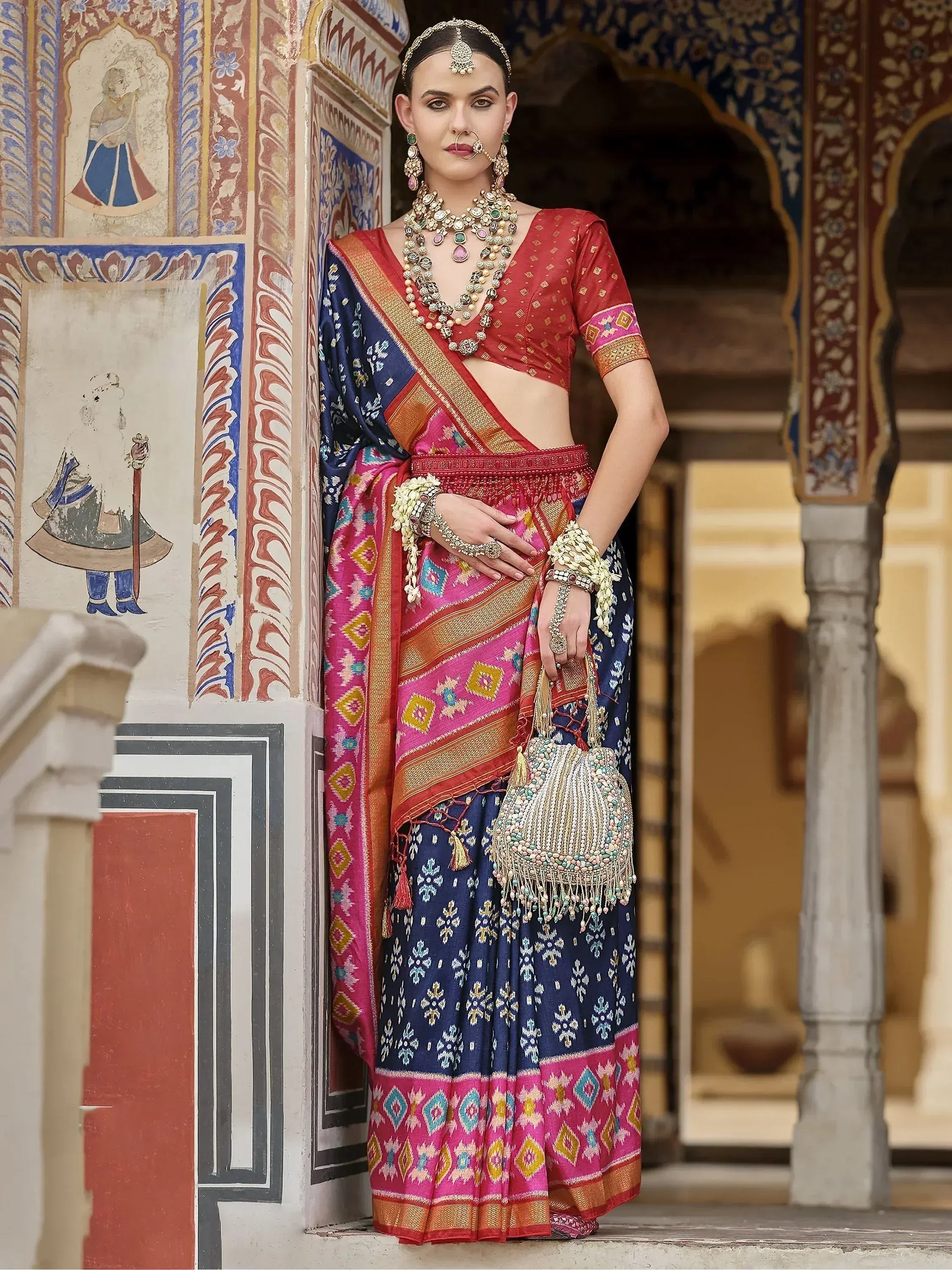 Woman in vibrant Indian ethnic saree with red blouse, blue and pink patterned skirt, traditional jewelry, standing near ornate palace wall