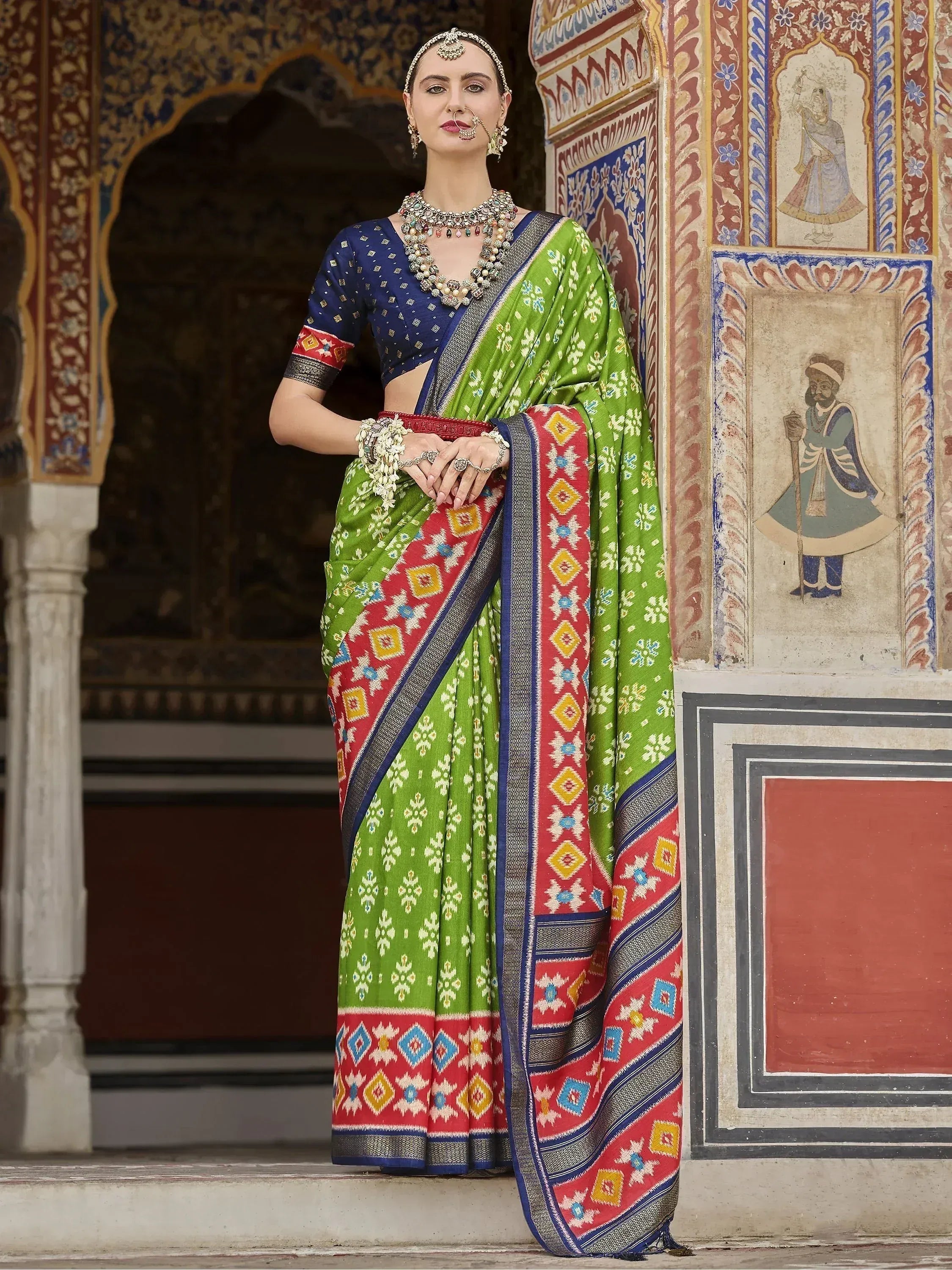 Woman in vibrant green and red printed saree with blue blouse, traditional Indian ethnic wear, standing by ornate palace wall.