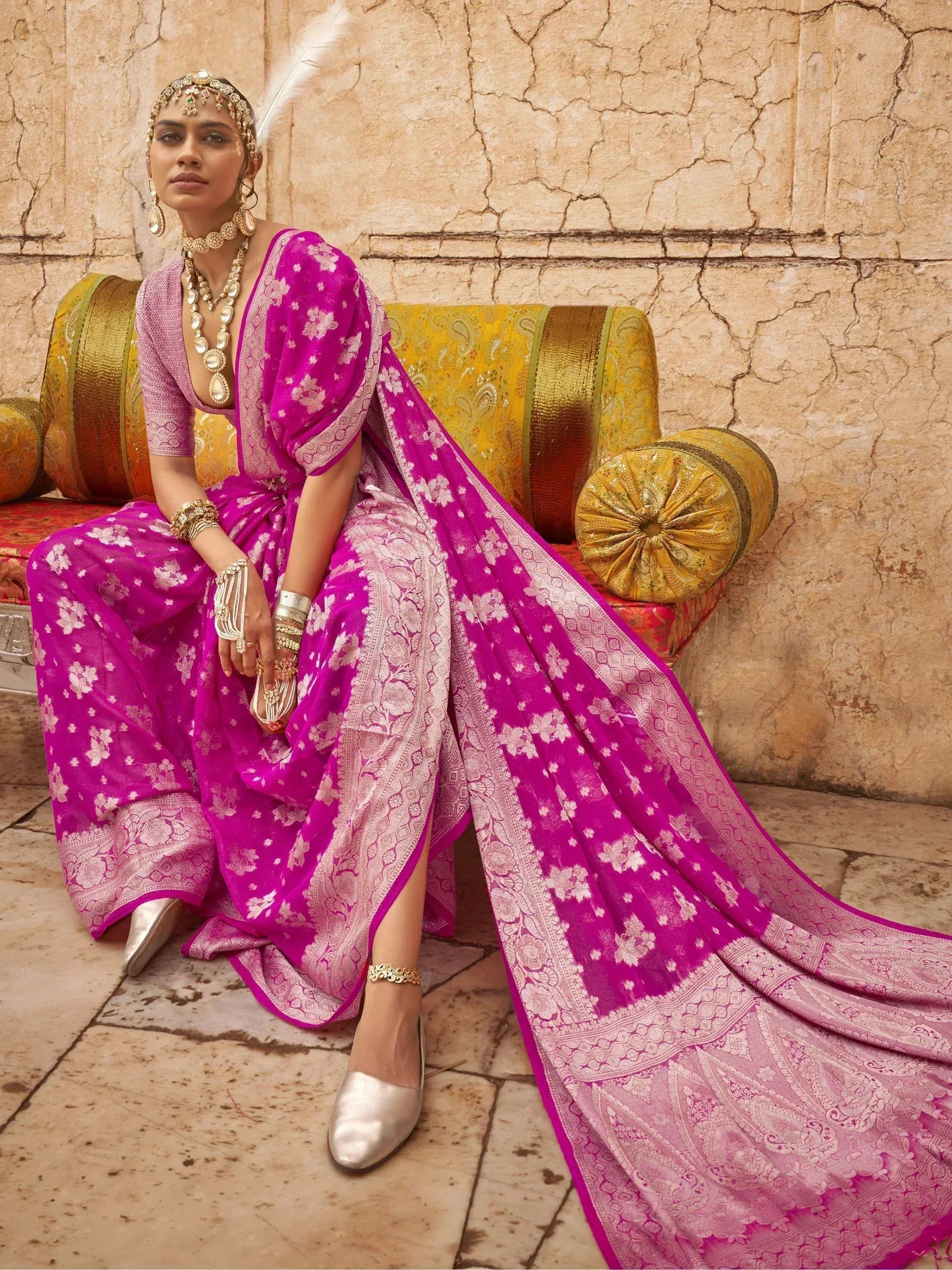 Woman in vibrant pink floral ethnic gown with gold jewelry, seated on ornate bench against stone wall