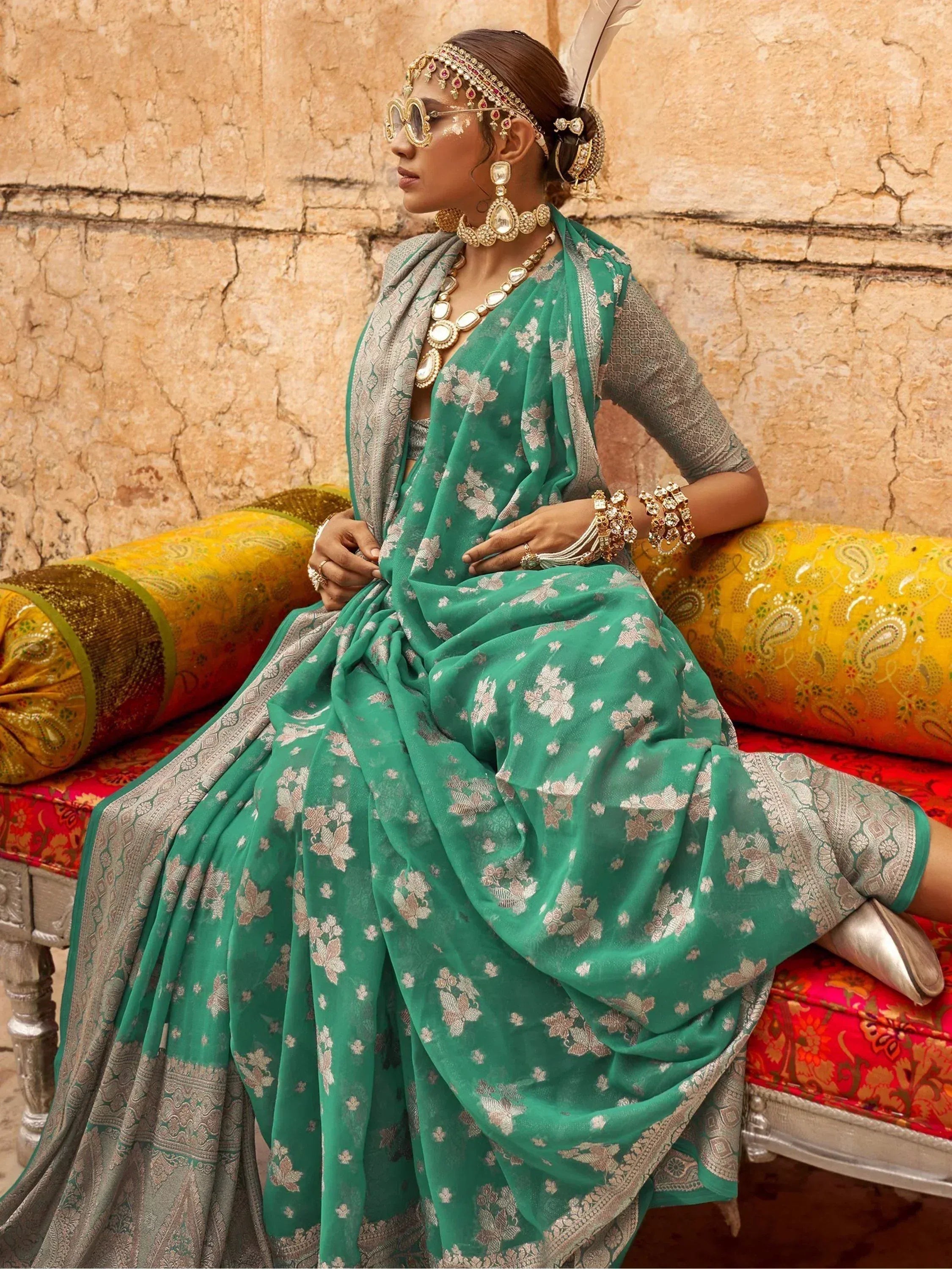 Woman in green floral saree with silver border, traditional Indian jewelry, and sunglasses, seated on ornate bench.