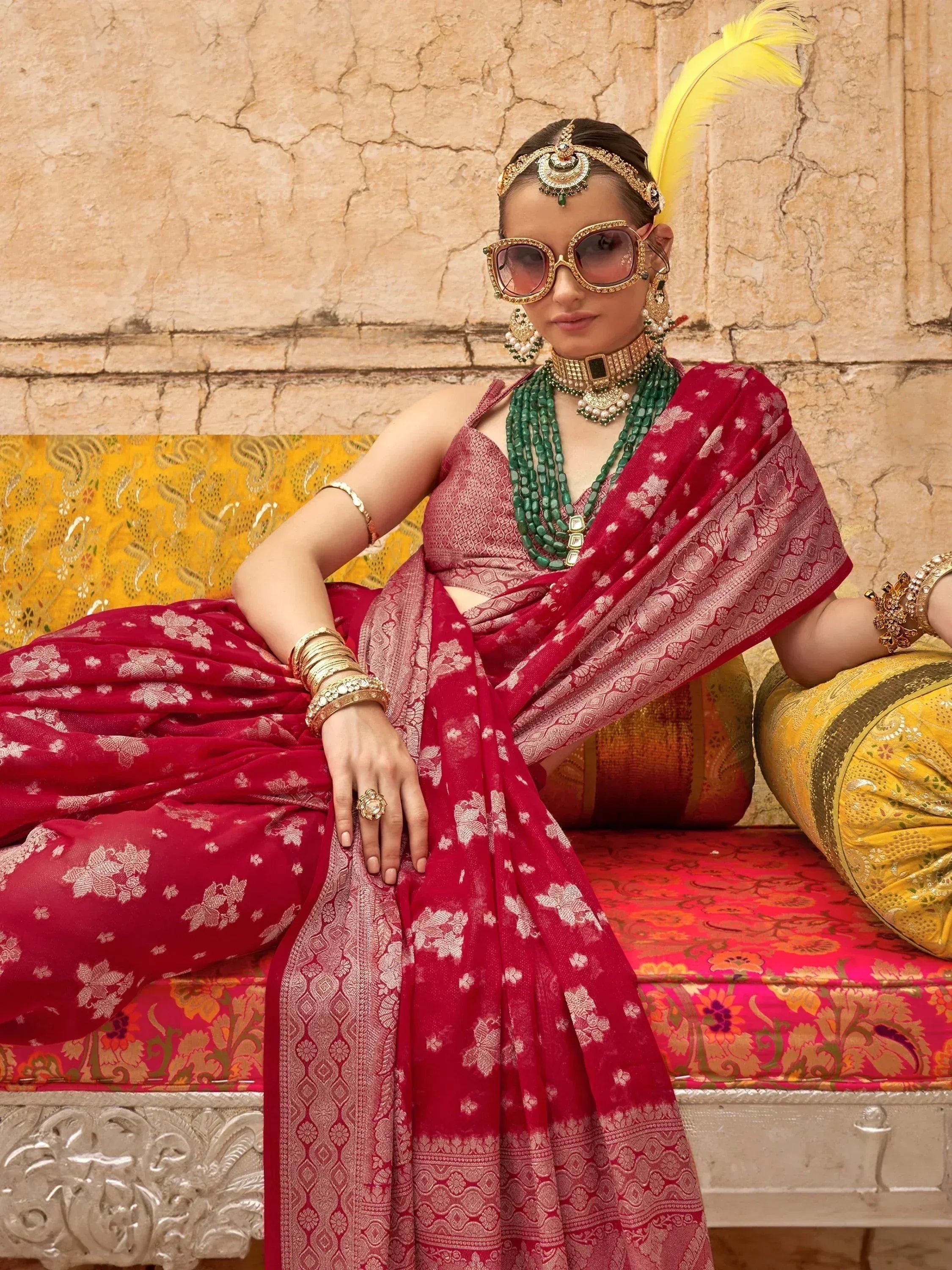 Woman in red floral saree with gold jewelry and sunglasses, seated on ornate bench