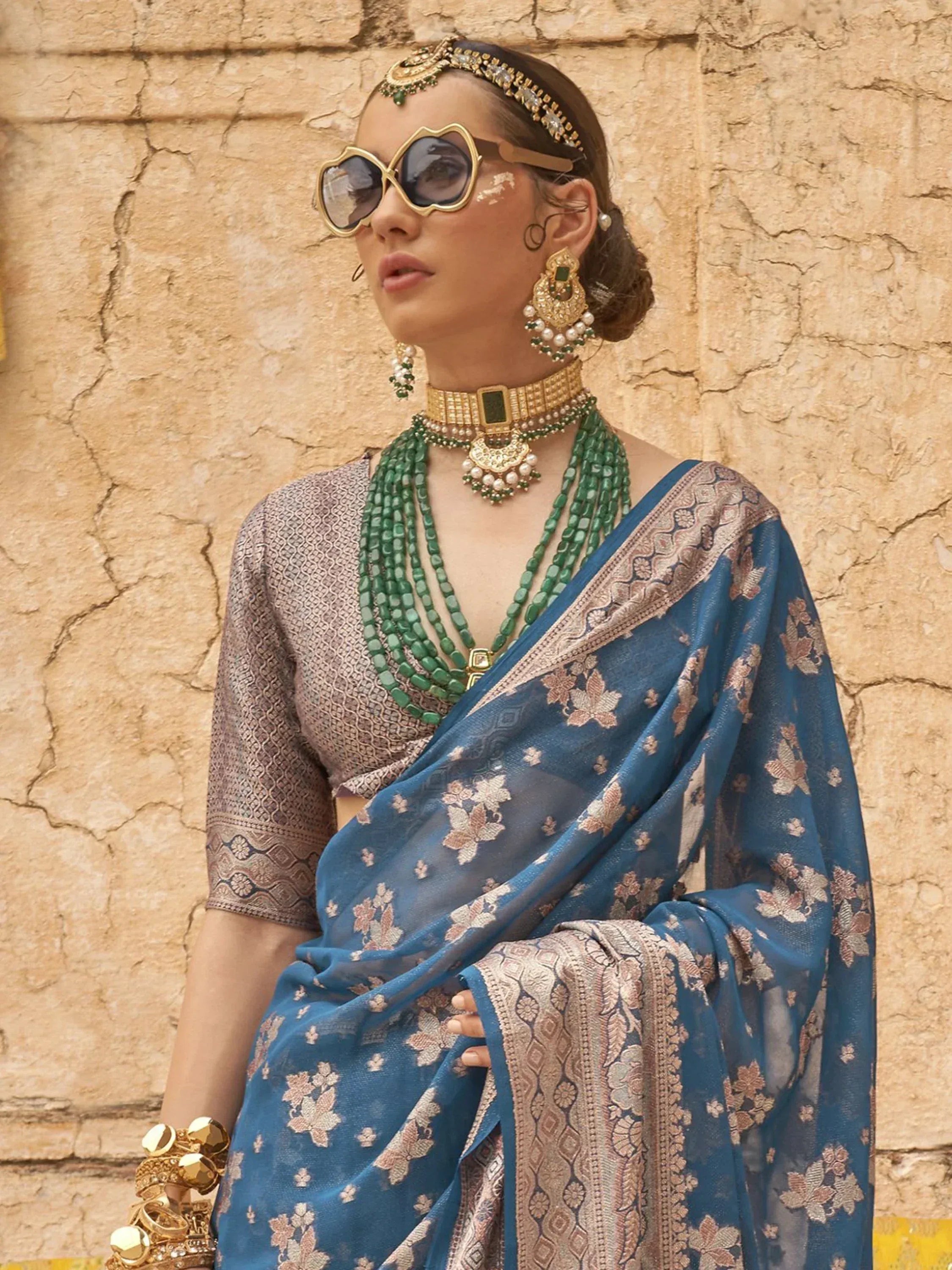 Woman in blue floral saree with gold embroidery, green jewelry, and statement sunglasses against stone wall