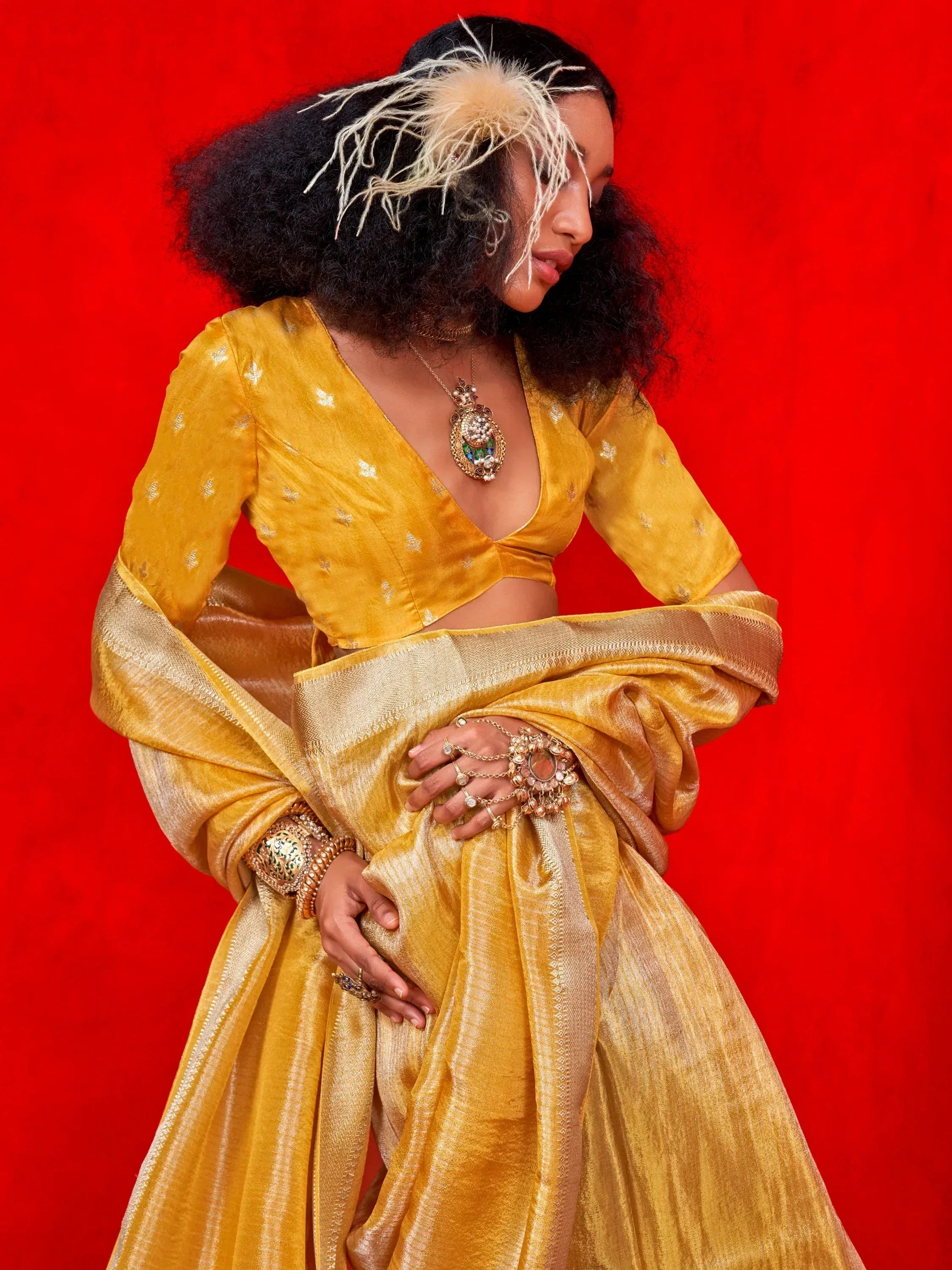 Golden ethnic saree with velvet blouse, intricate jewelry, and feather hair accessory against red backdrop