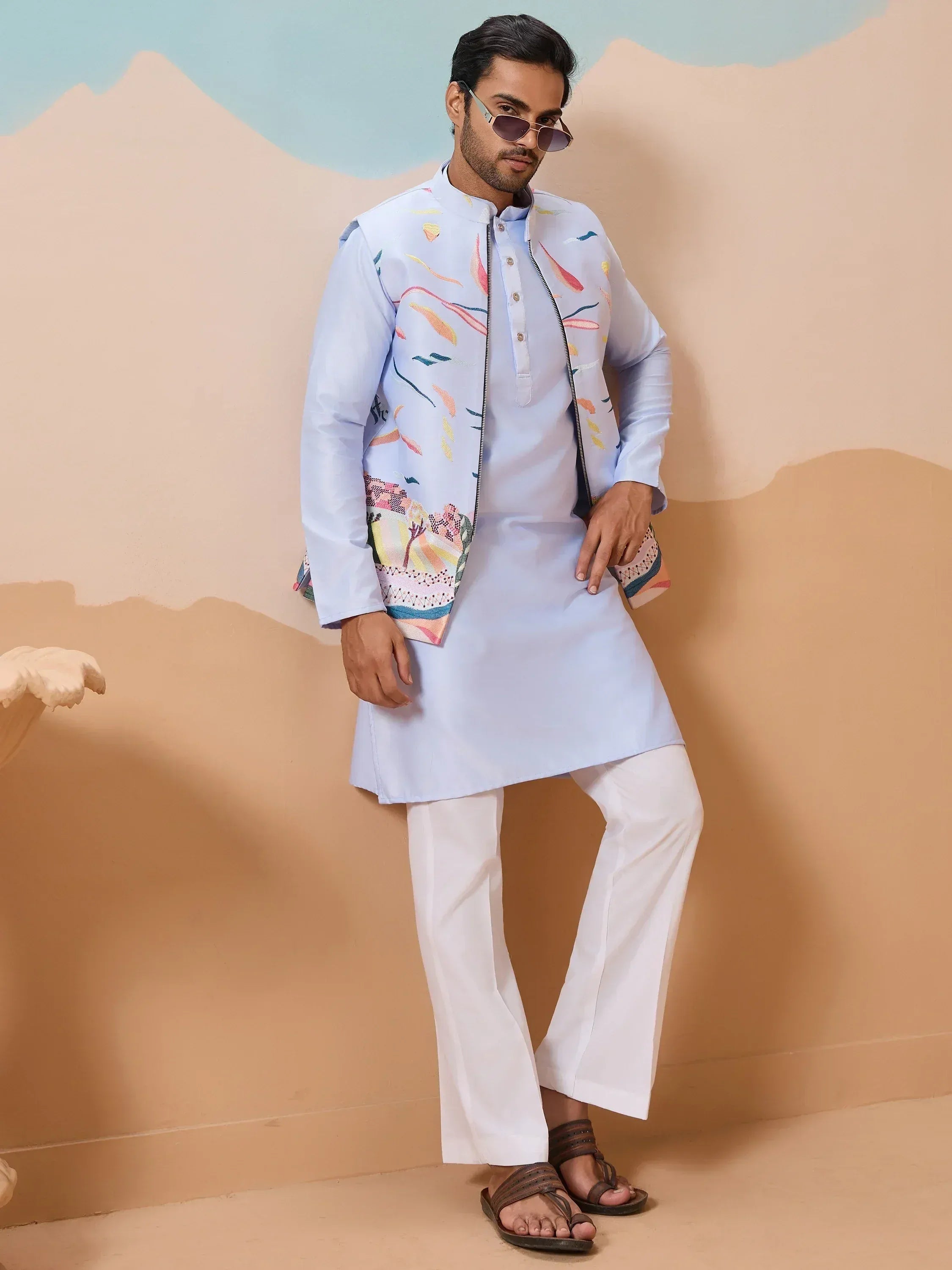 Man in light blue kurta with colorful embroidered jacket, white pants, and sandals for ethnic wear