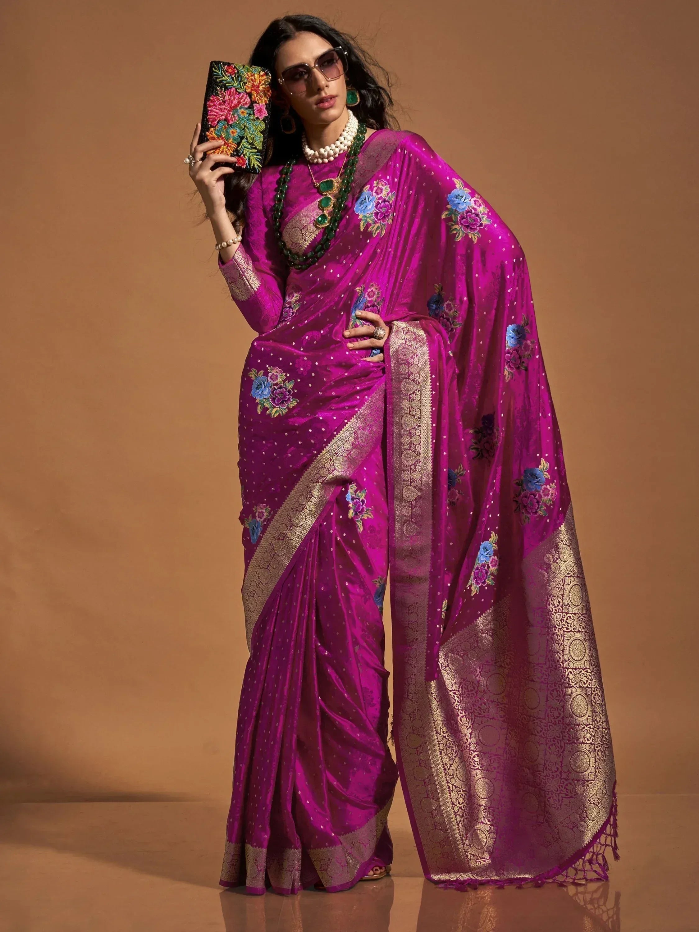 Vibrant magenta saree with floral embroidery and gold border, styled with statement jewelry and clutch.
