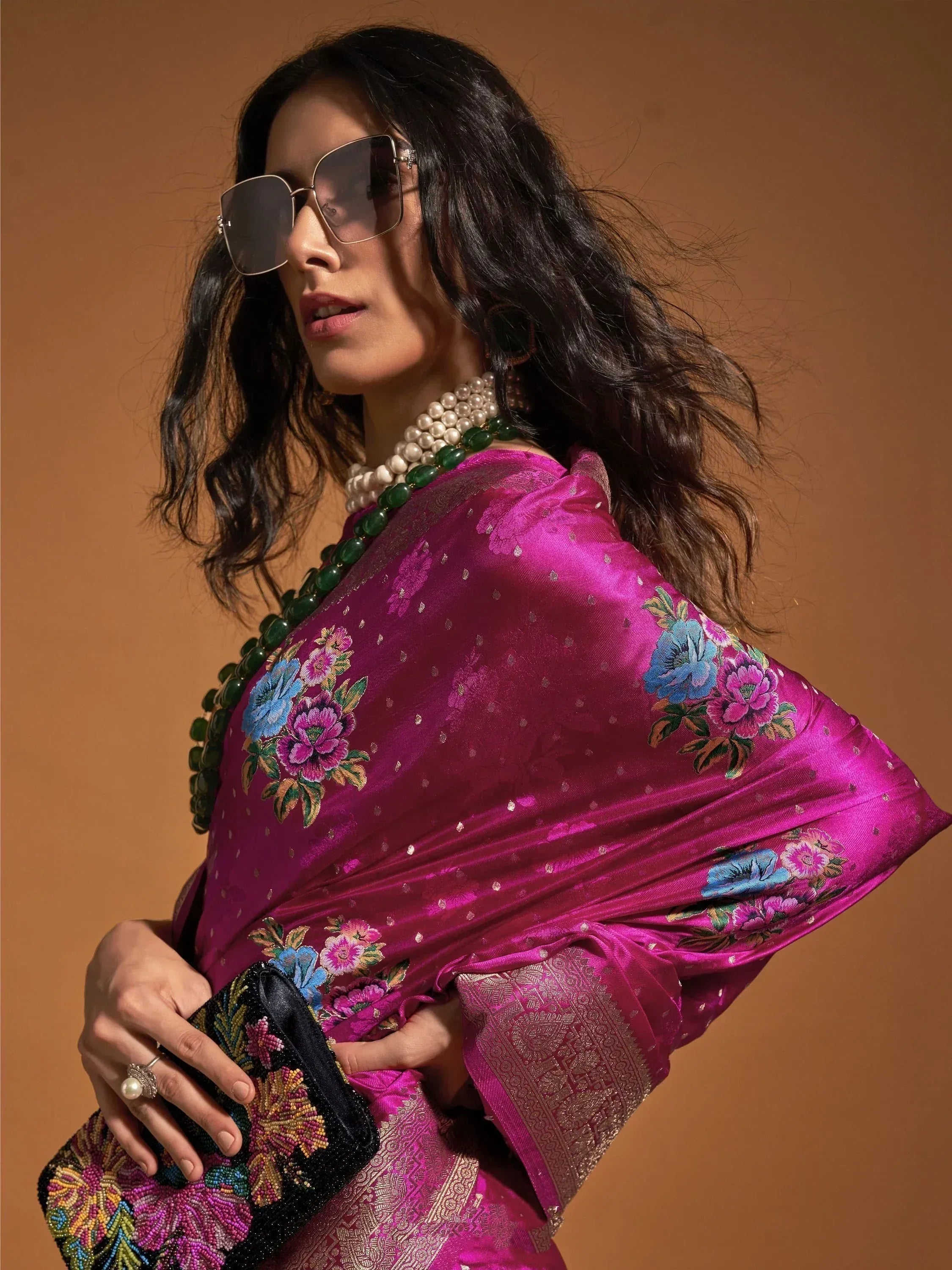 Woman in magenta floral embroidered saree with pearl and green bead necklace, holding beaded clutch