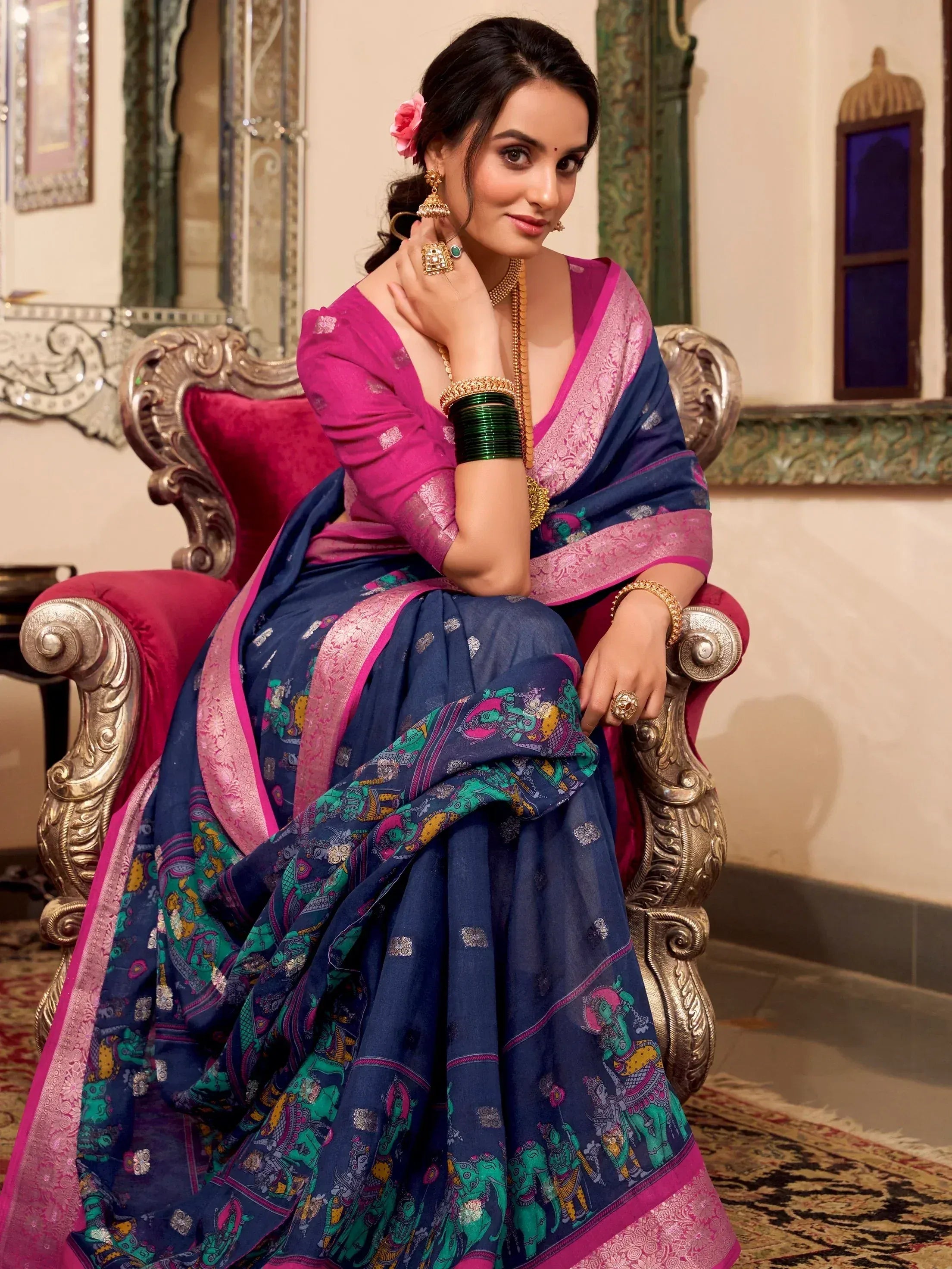Woman in navy blue and pink printed saree with gold jewelry, seated on ornate chair
