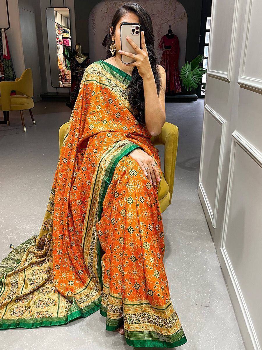 Woman in vibrant orange printed saree with green border, seated in boutique, capturing mirror selfie