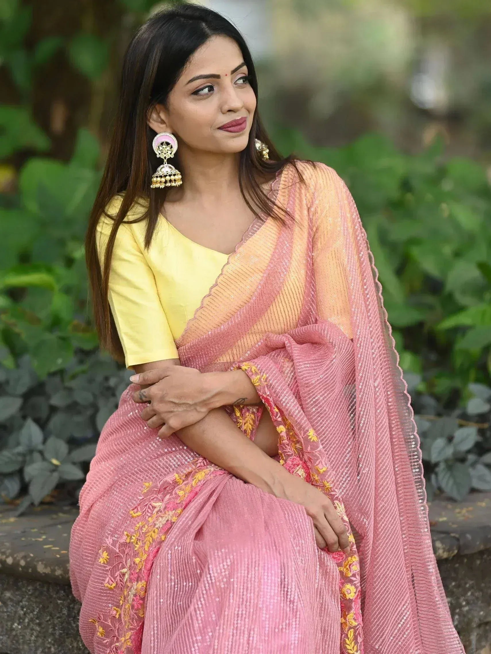 Woman in pink sequin saree with yellow blouse, floral embroidery, and jhumka earrings, outdoor garden setting