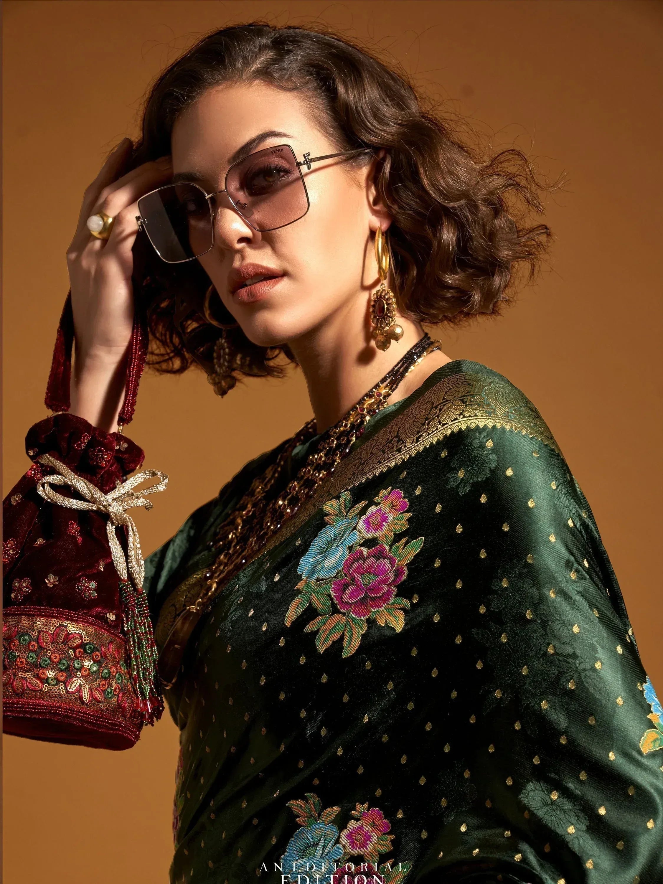 Woman in green embroidered saree with floral motifs, gold jewelry, and sunglasses, holding a red velvet potli bag.