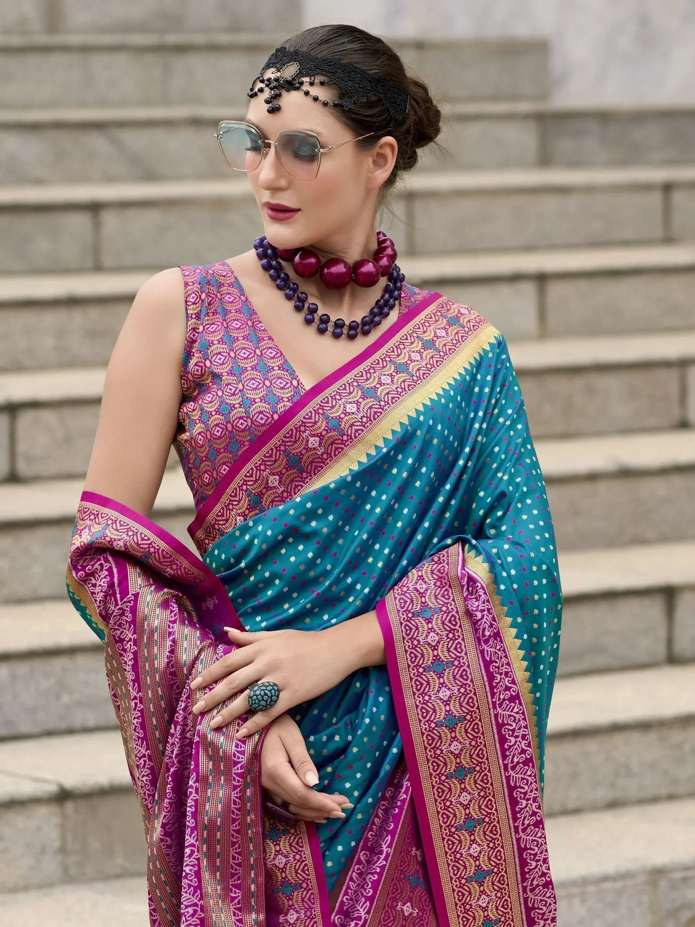 Woman in vibrant teal and magenta ethnic saree with intricate patterns, styled with statement jewelry and sunglasses.