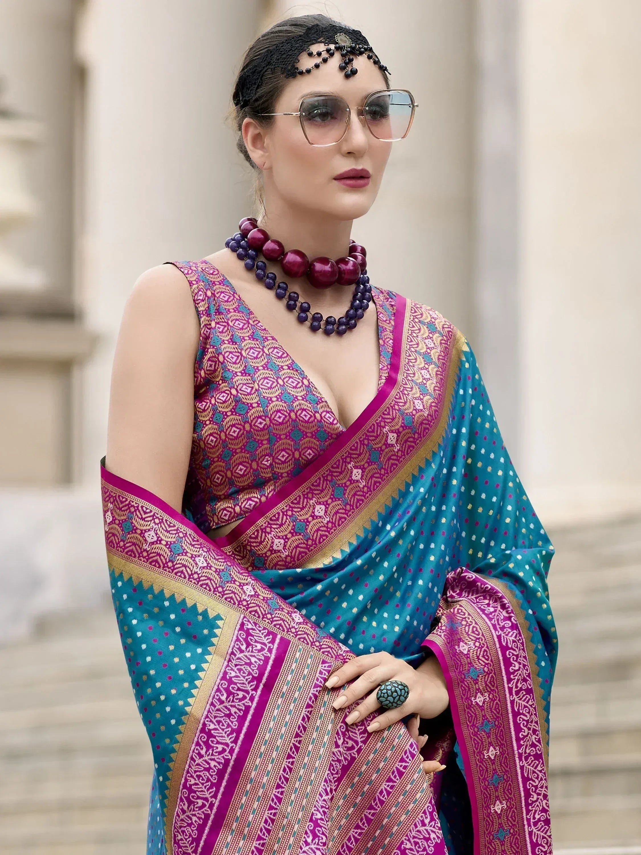 Woman in vibrant purple and teal designer saree with intricate patterns, ethnic jewelry, and sunglasses
