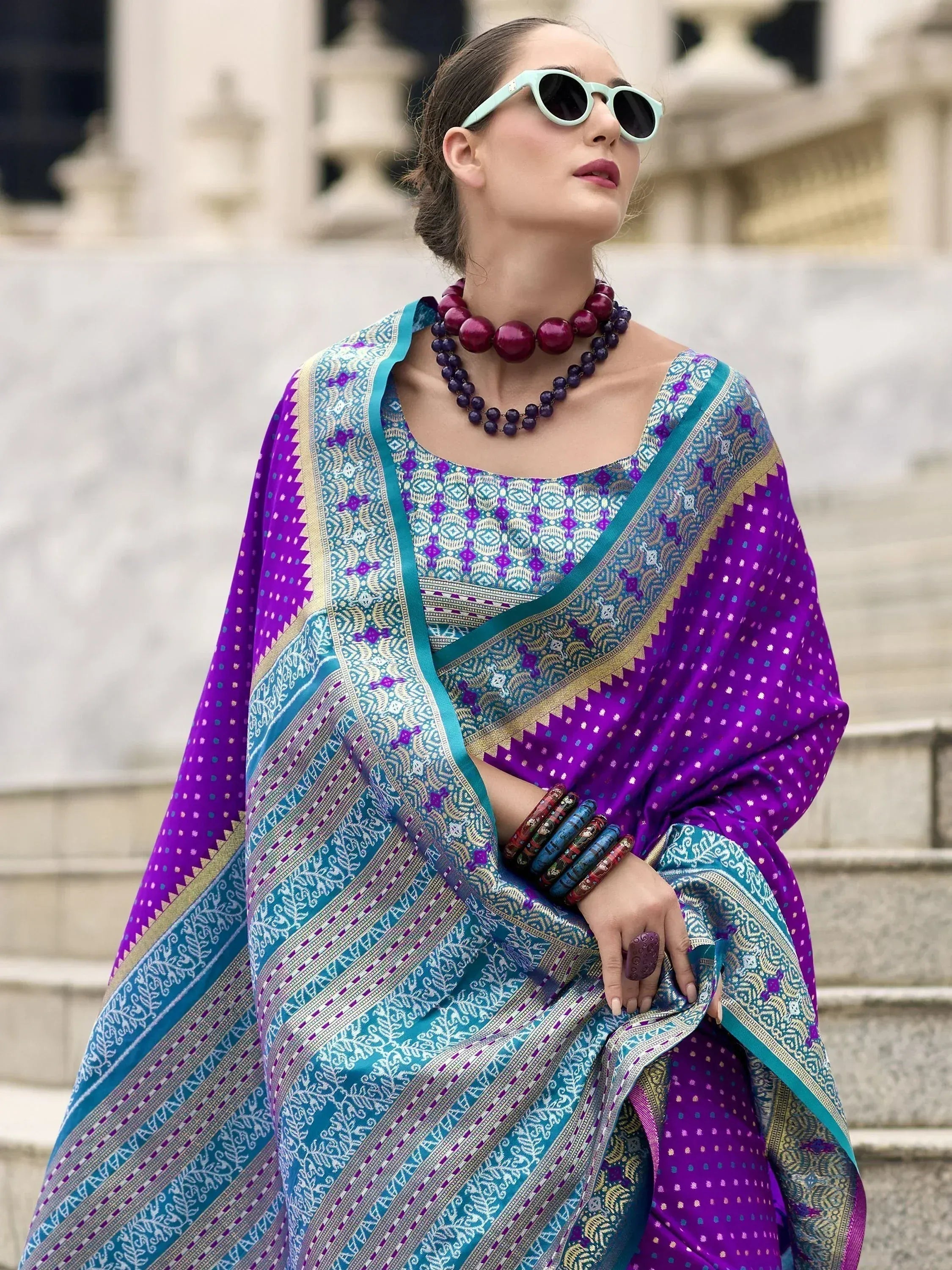 Woman in vibrant purple and teal ethnic saree with intricate patterns, styled with bold jewelry and sunglasses.