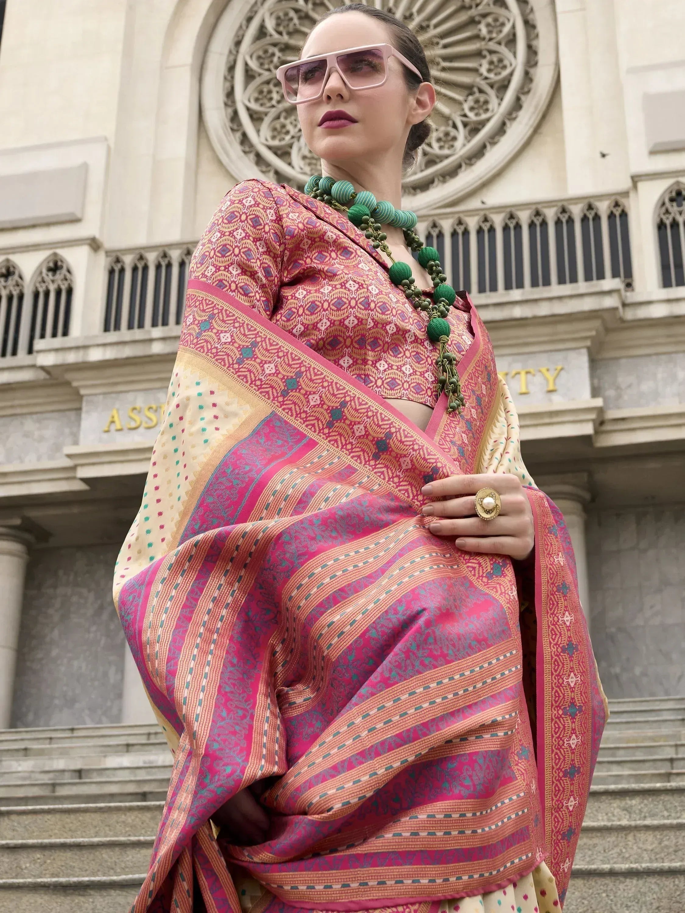 Woman in vibrant pink and gold patterned Indian saree with green necklace, standing before ornate building
