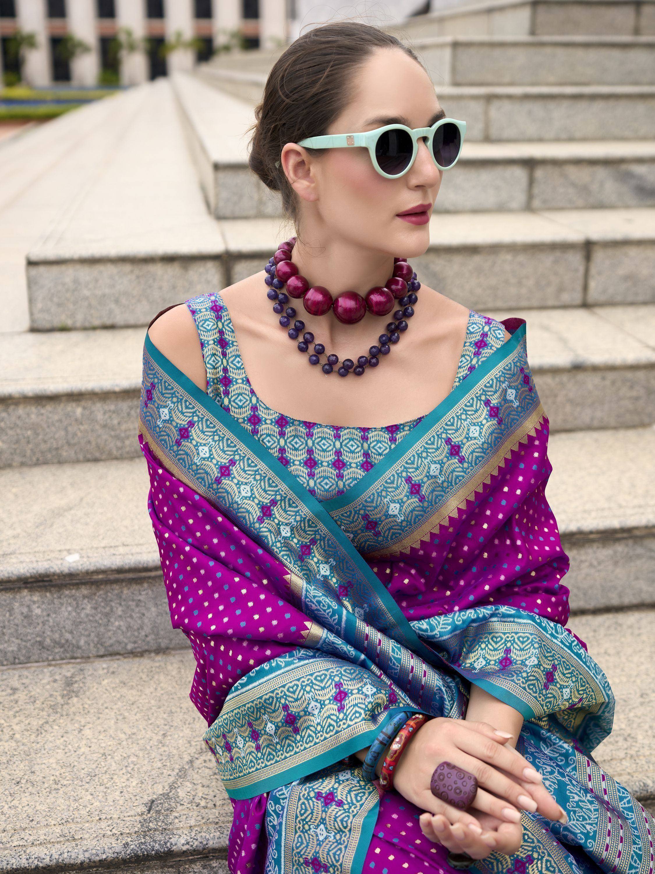 Woman in vibrant purple and teal designer saree with gold patterns, styled with statement necklace and sunglasses