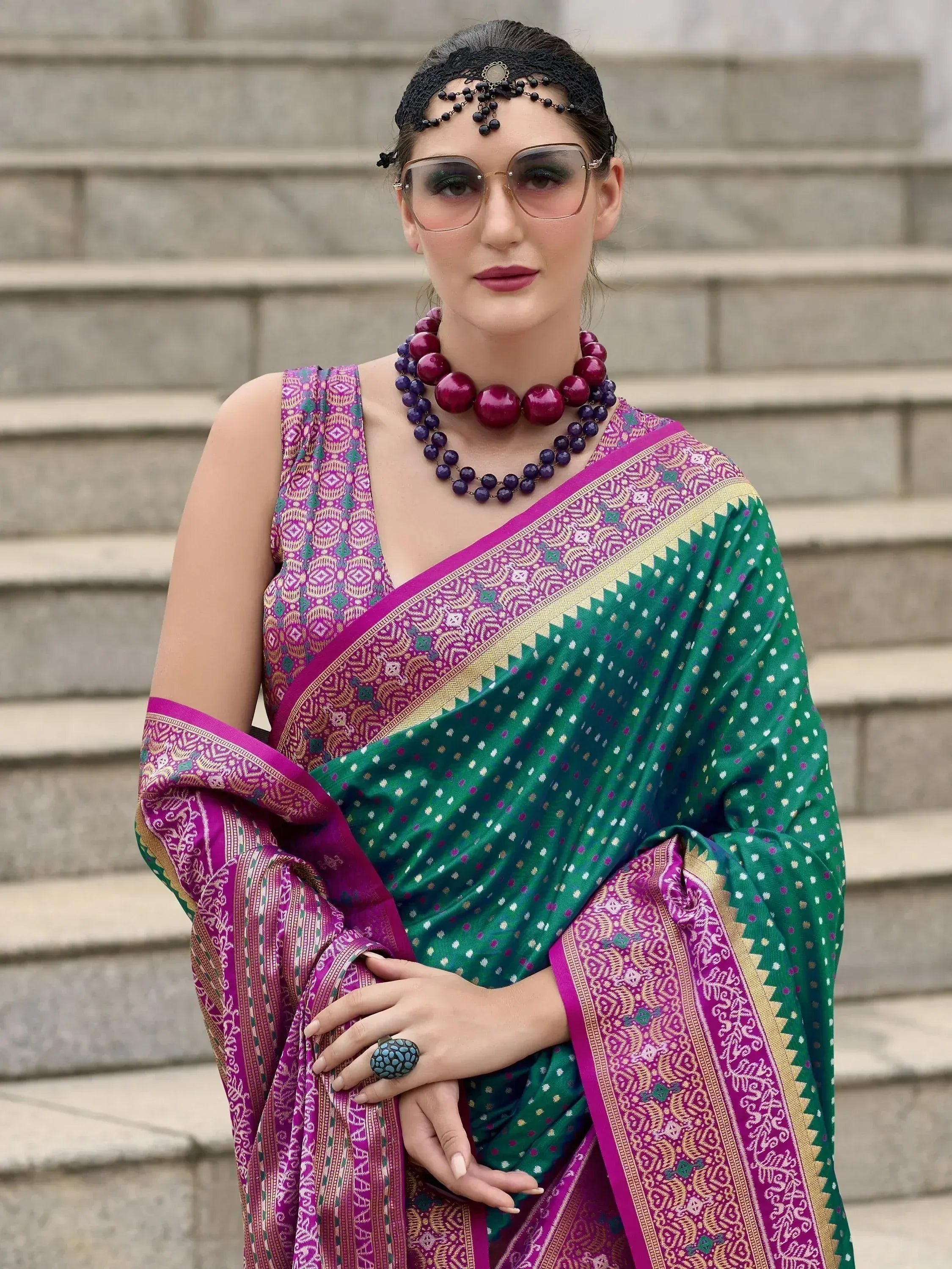 Woman in vibrant green and purple ethnic saree with intricate patterns, styled with bold jewelry and sunglasses.