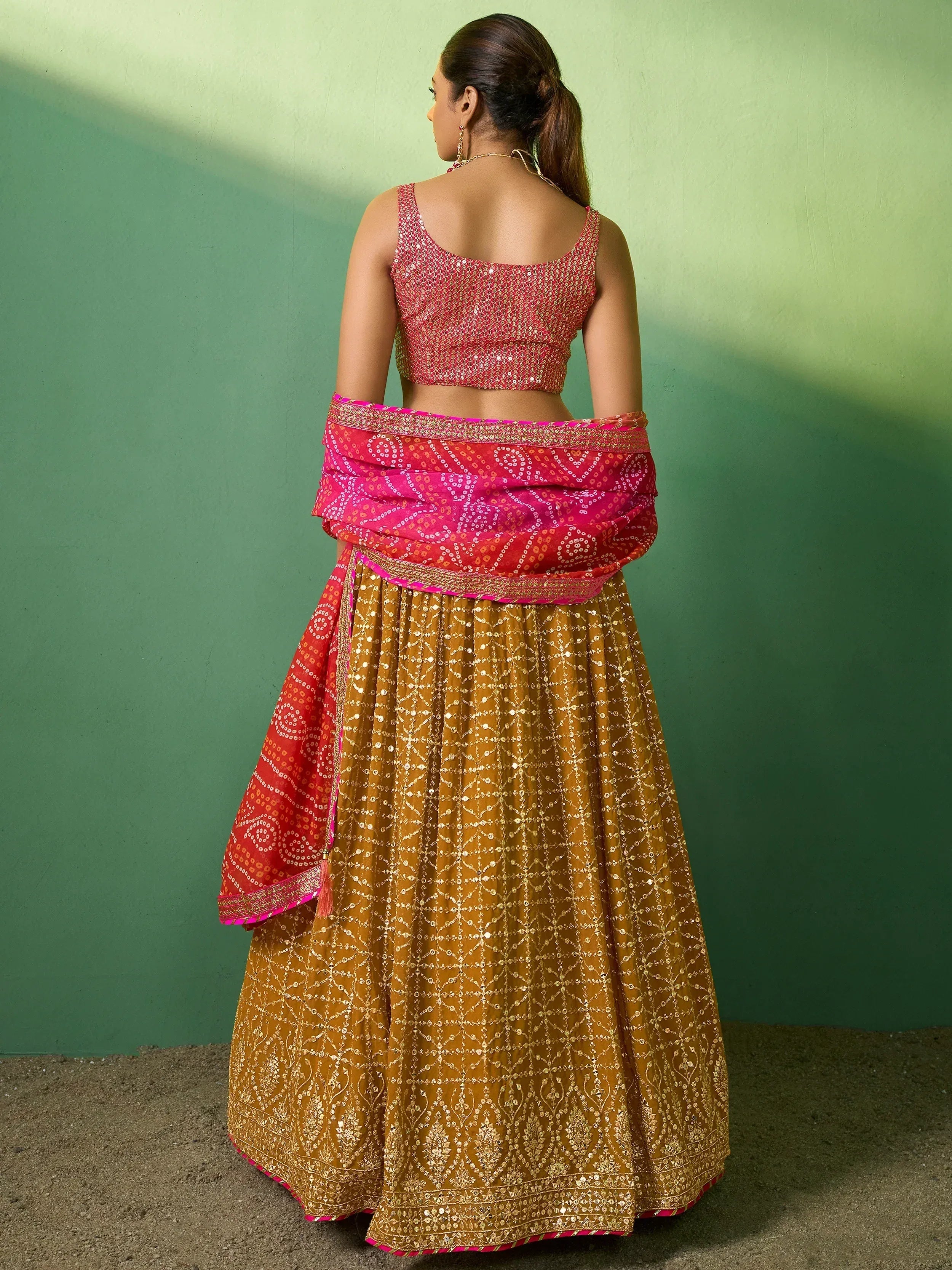 Woman in pink and gold designer lehenga choli with sequin work, back view against green wall