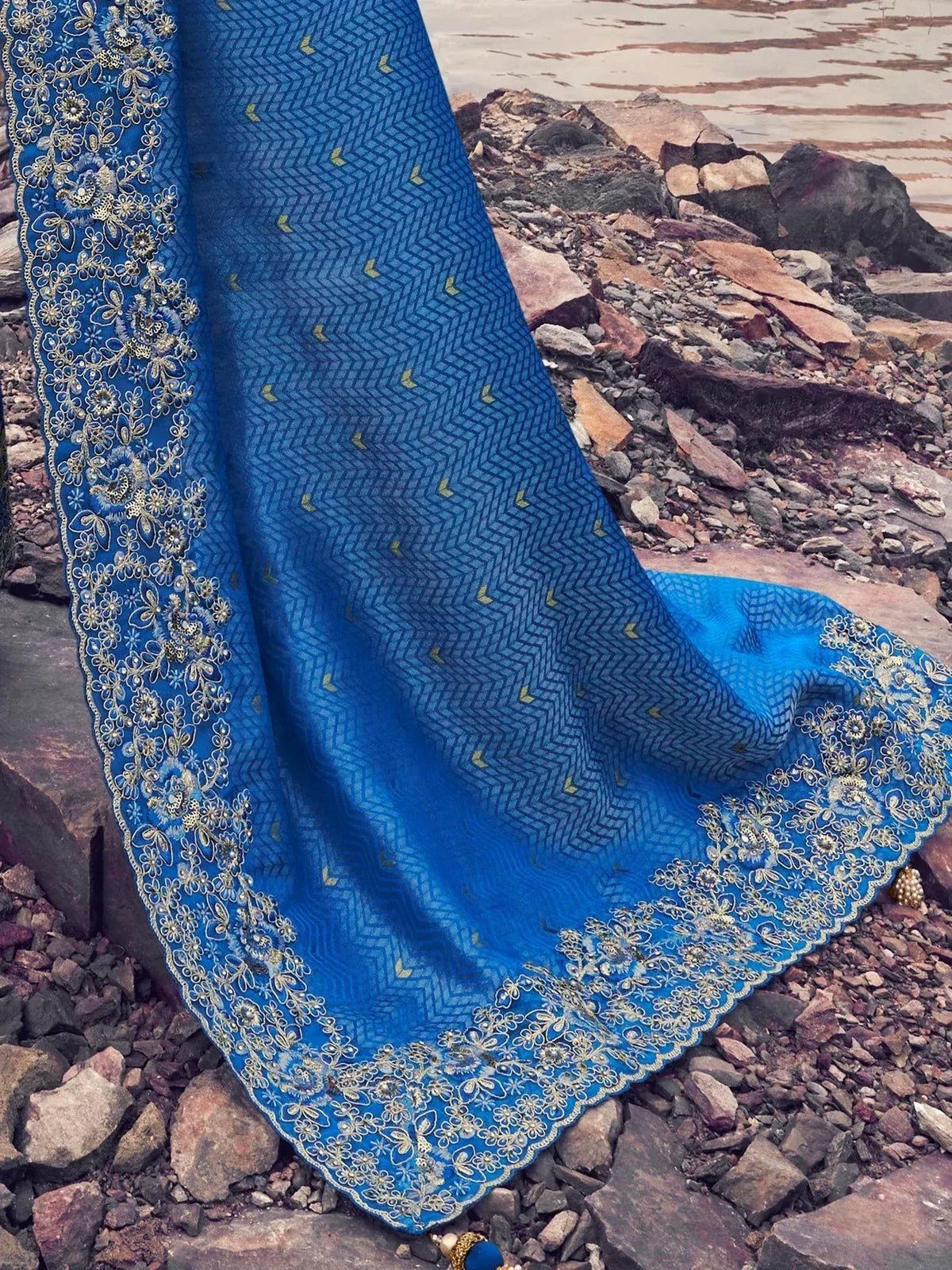 Blue embroidered saree with gold floral border, herringbone pattern, displayed on rocky shore