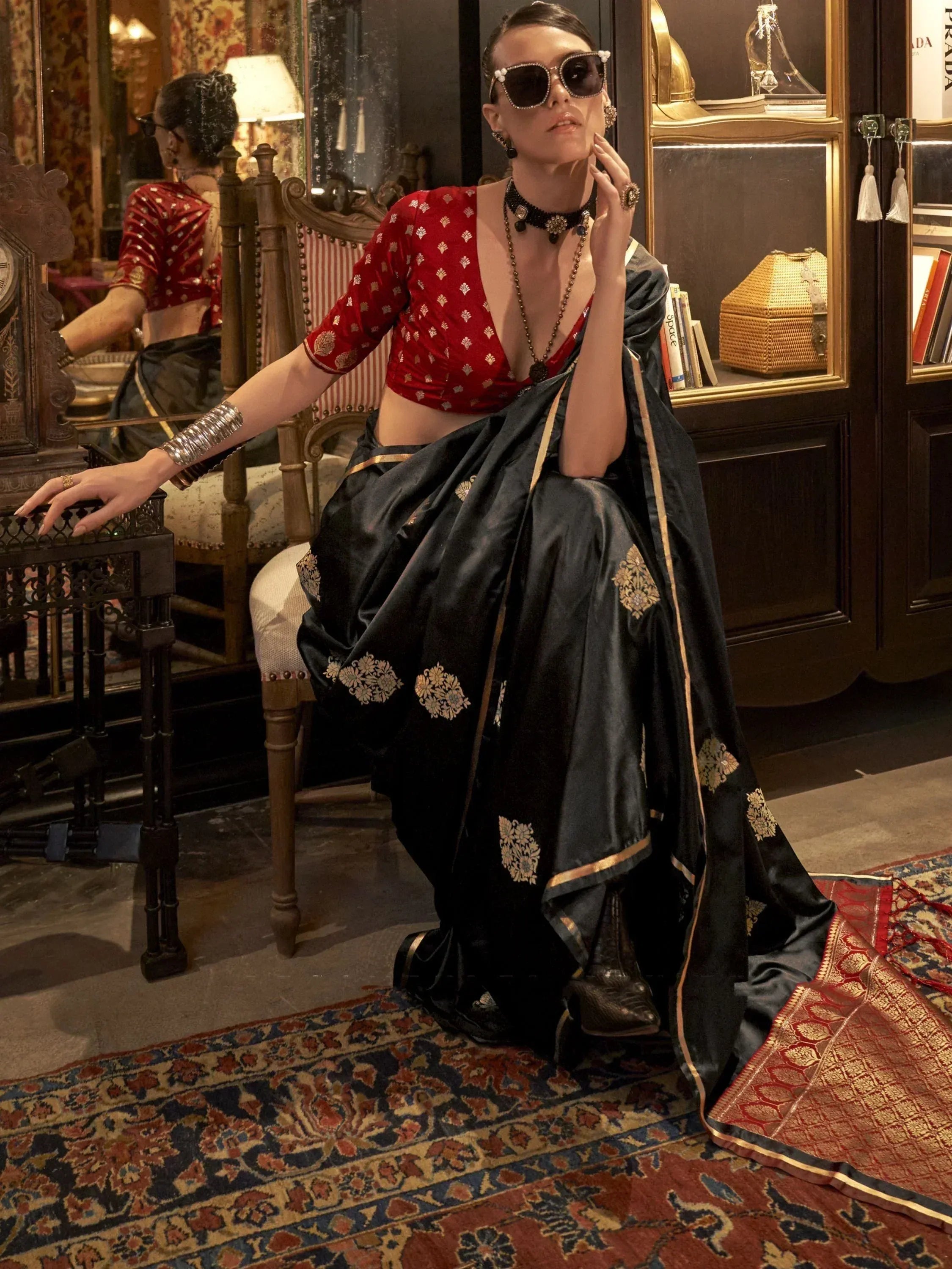 Woman in black silk saree with gold embroidery and red blouse, styled with statement jewelry and sunglasses.