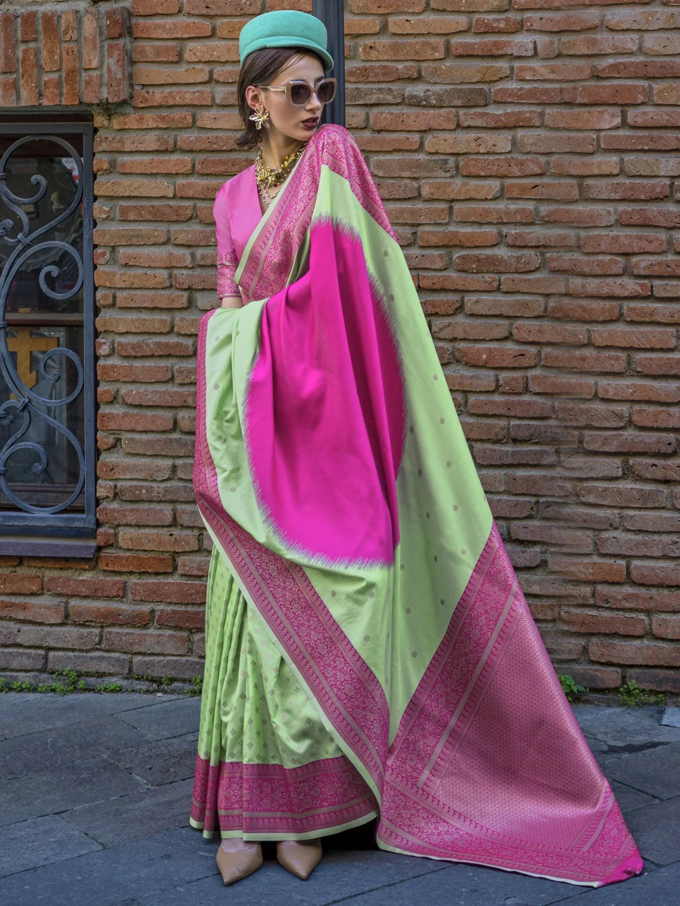 Woman in vibrant pink and green designer saree with gold jewelry, standing against brick wall