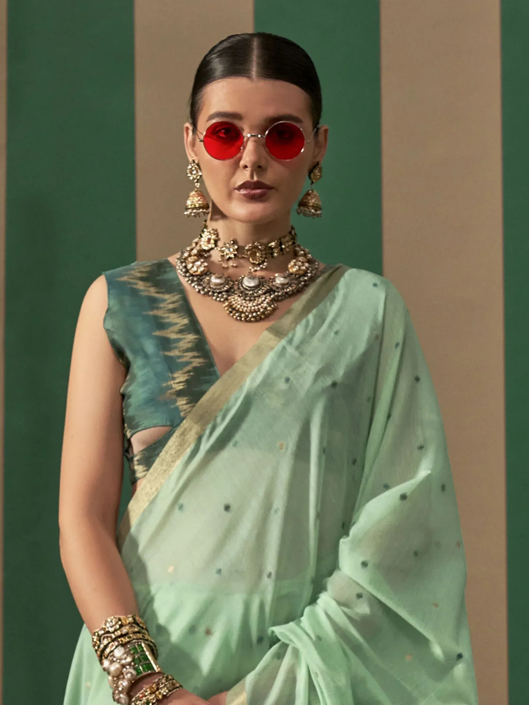 Woman in mint green saree with gold blouse, statement jewelry, and red sunglasses, styled for ethnic wear.