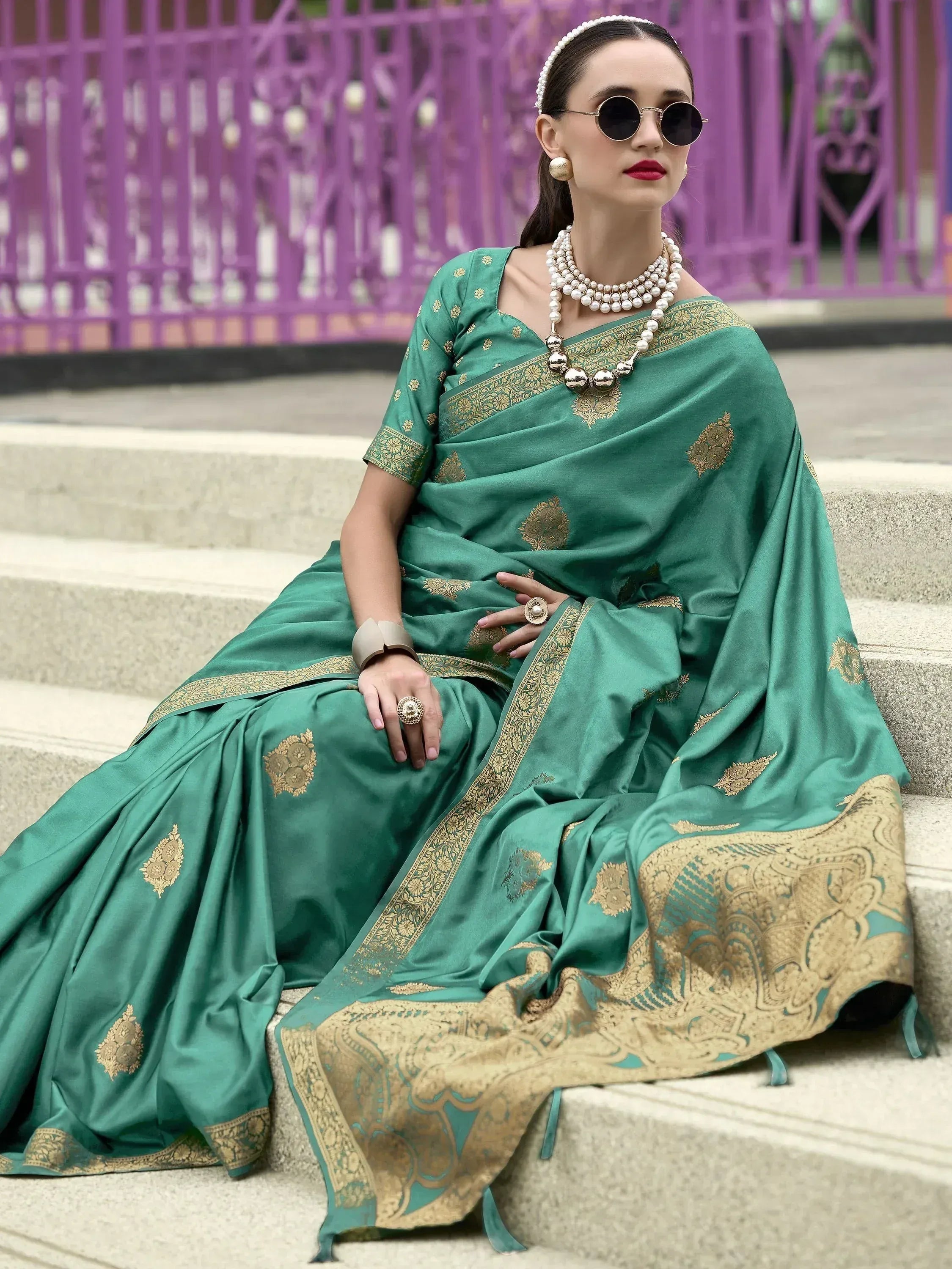 Woman in green silk saree with gold embroidery, styled with pearl jewelry and sunglasses, sitting on steps.