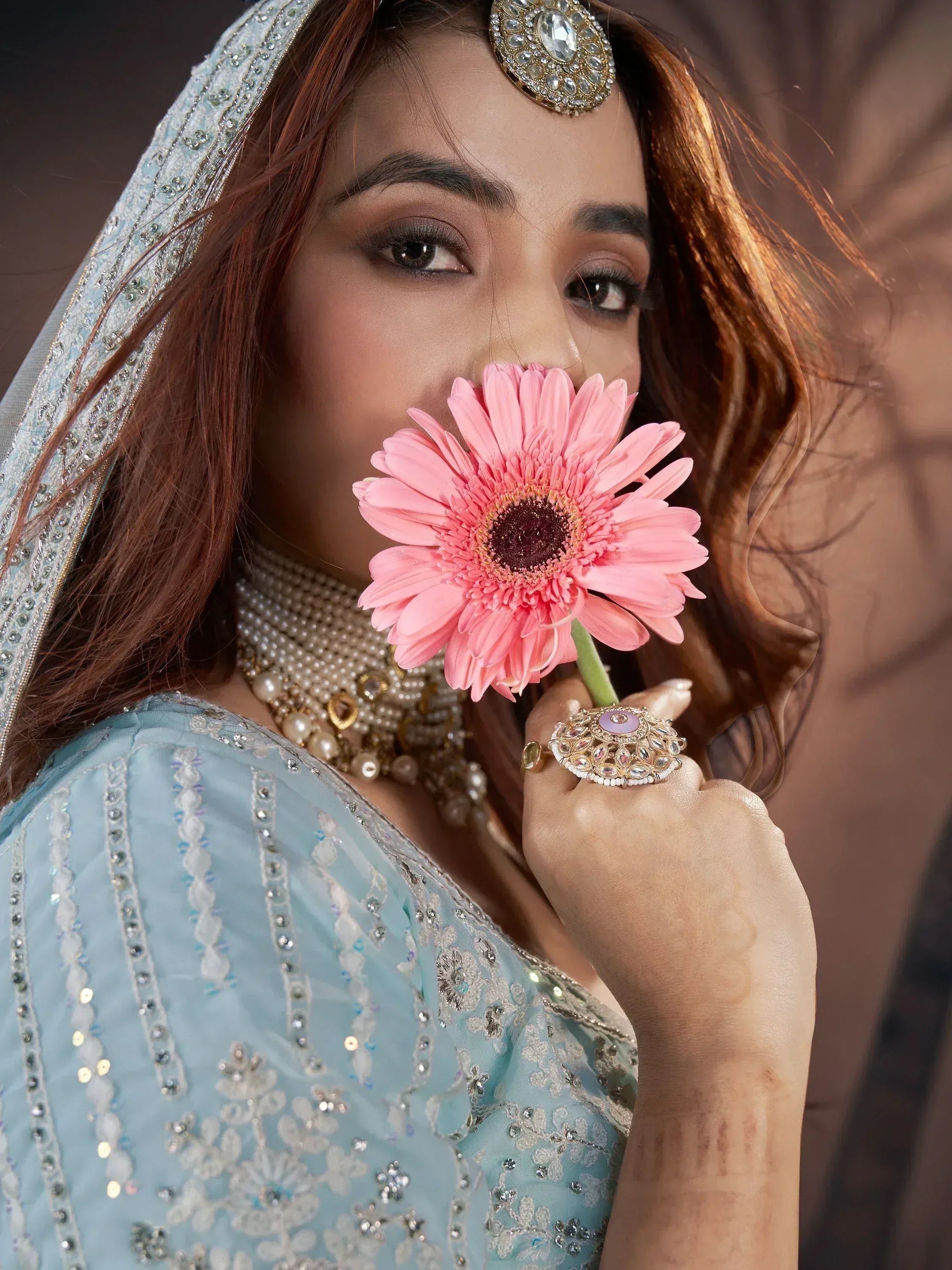 Woman in light blue embroidered ethnic gown with pearl jewelry, holding pink flower, wearing maang tikka
