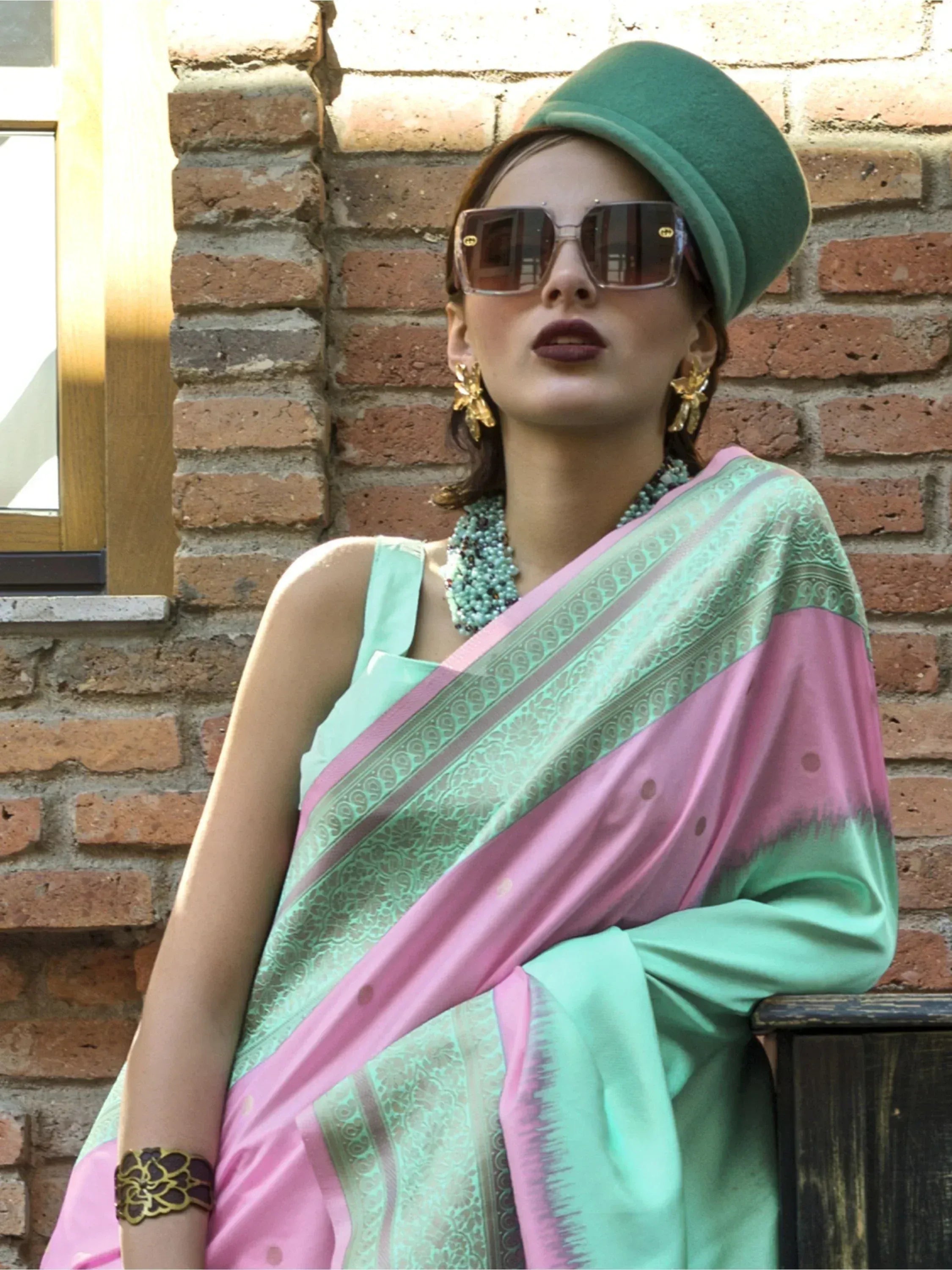 Stylish woman in pink and mint saree with green hat, gold jewelry, and sunglasses against brick wall
