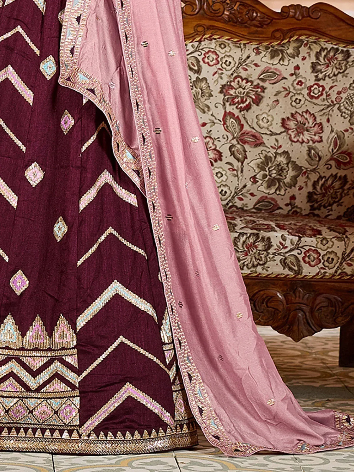 Maroon ethnic gown with geometric embroidery and pink dupatta, styled near floral sofa