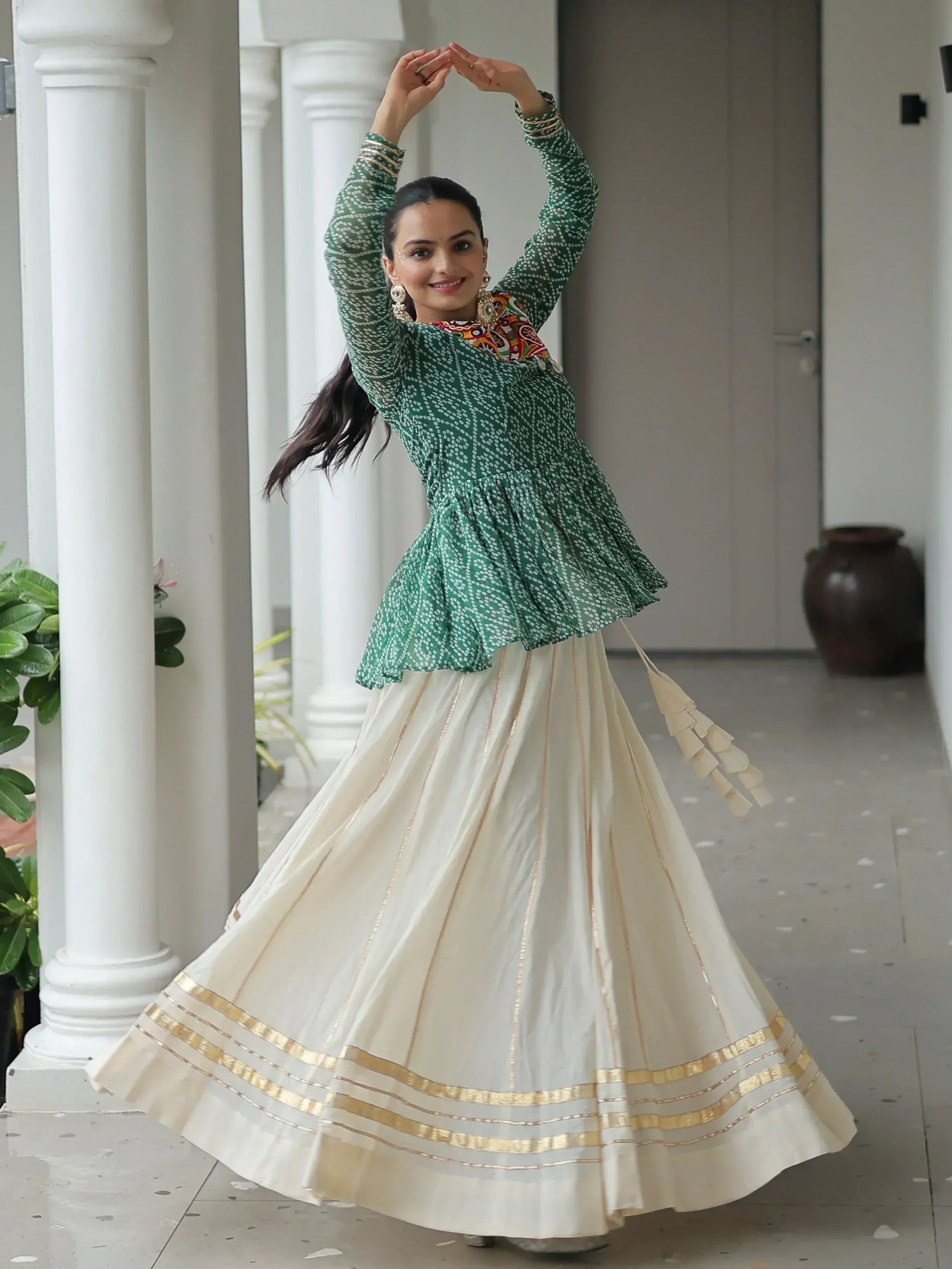 Woman in green printed top and cream pleated skirt with gold trim, dancing near white pillars