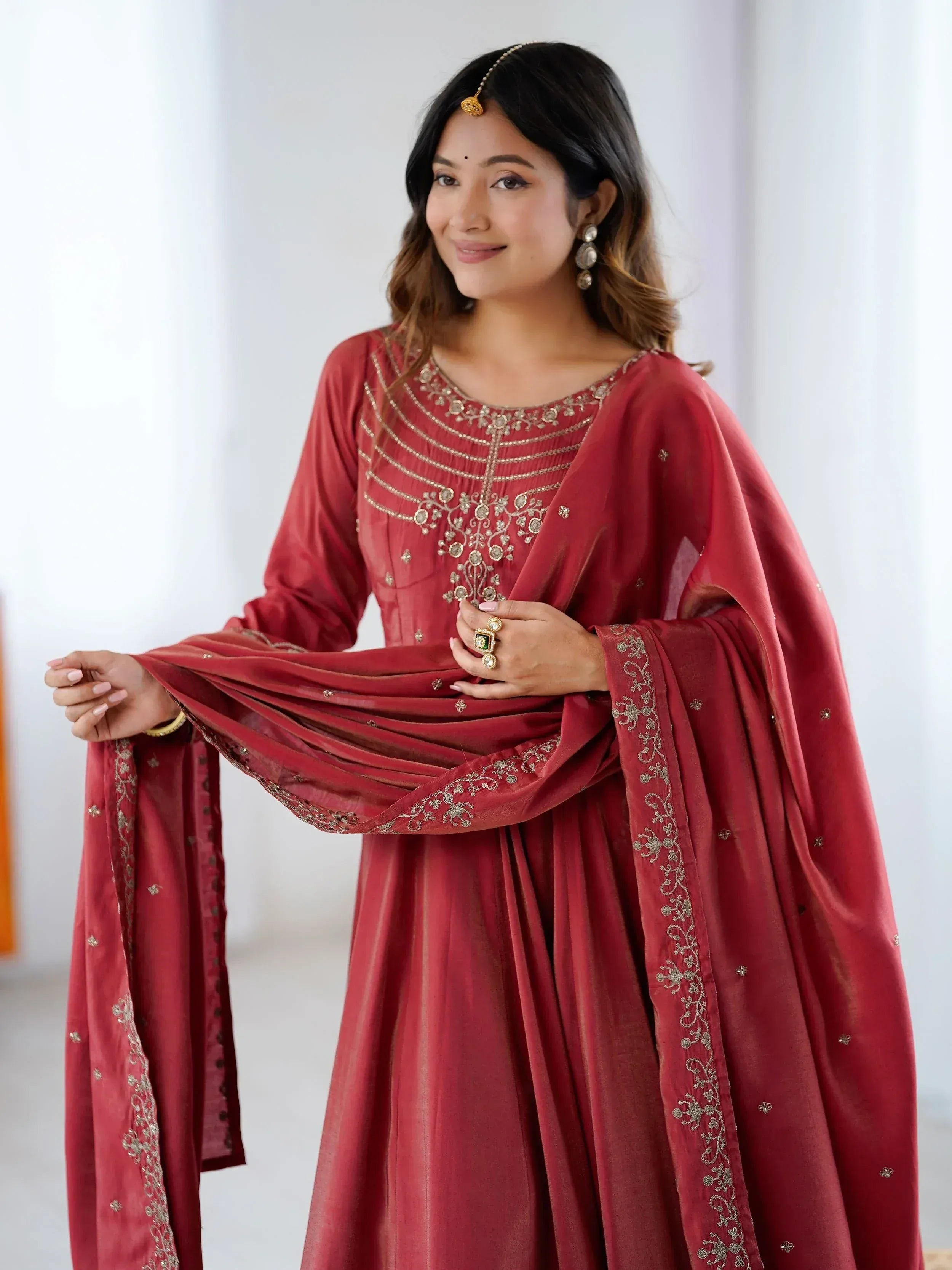 Woman in maroon embroidered salwar kameez with gold detailing and matching dupatta, styled with traditional jewelry.