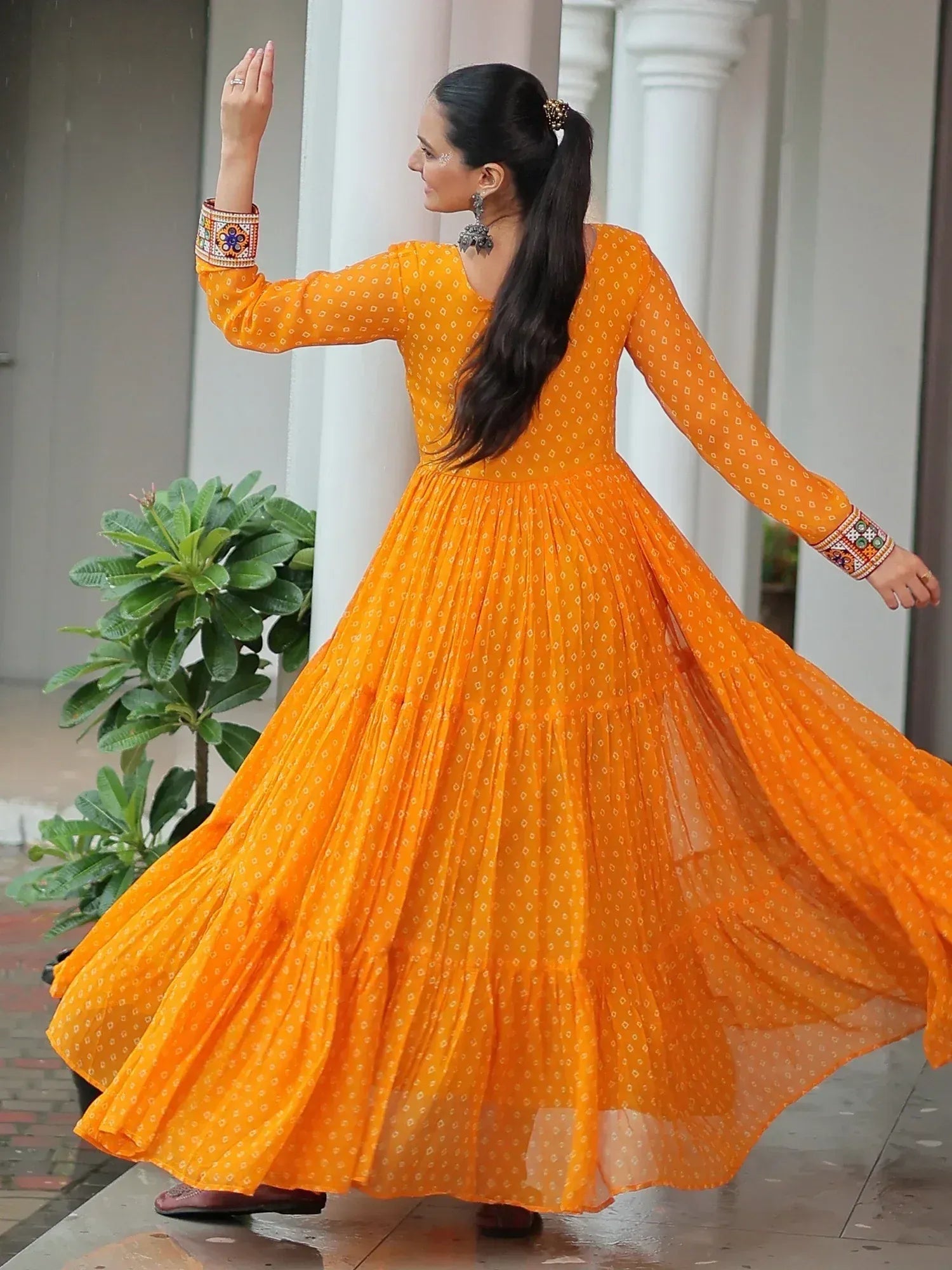 Woman twirling in vibrant orange ethnic gown with printed pattern and embroidered sleeves, perfect for festive occasions.