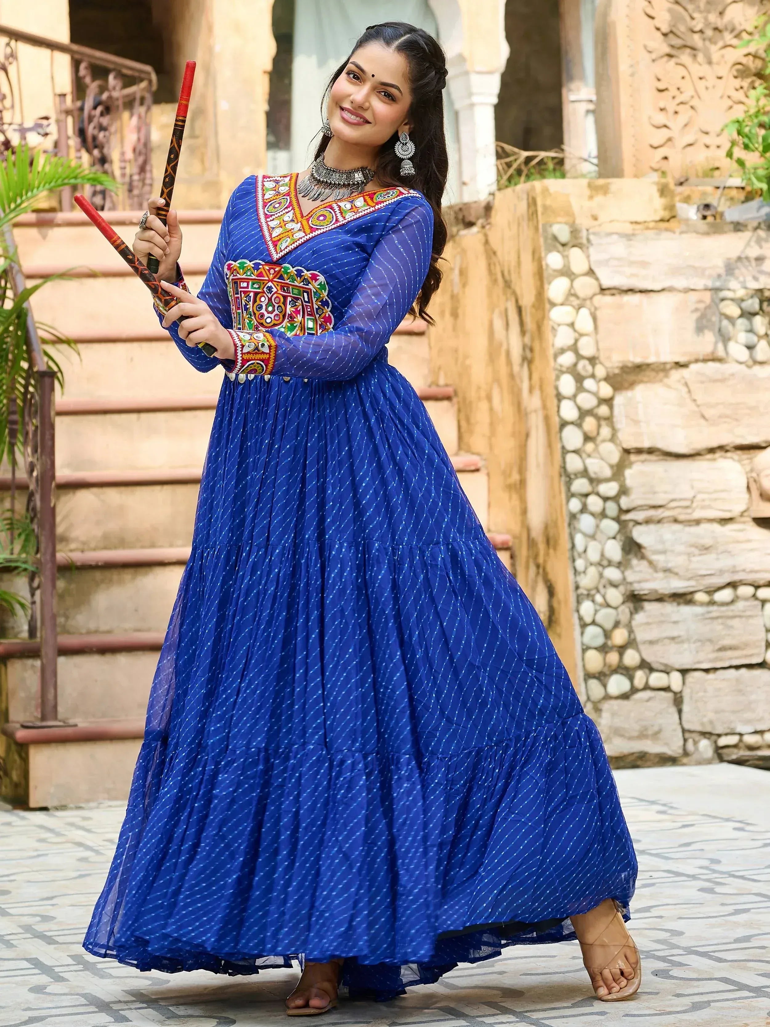 Woman in blue embroidered ethnic gown with mirror work, holding dandiya sticks, perfect for festive occasions.