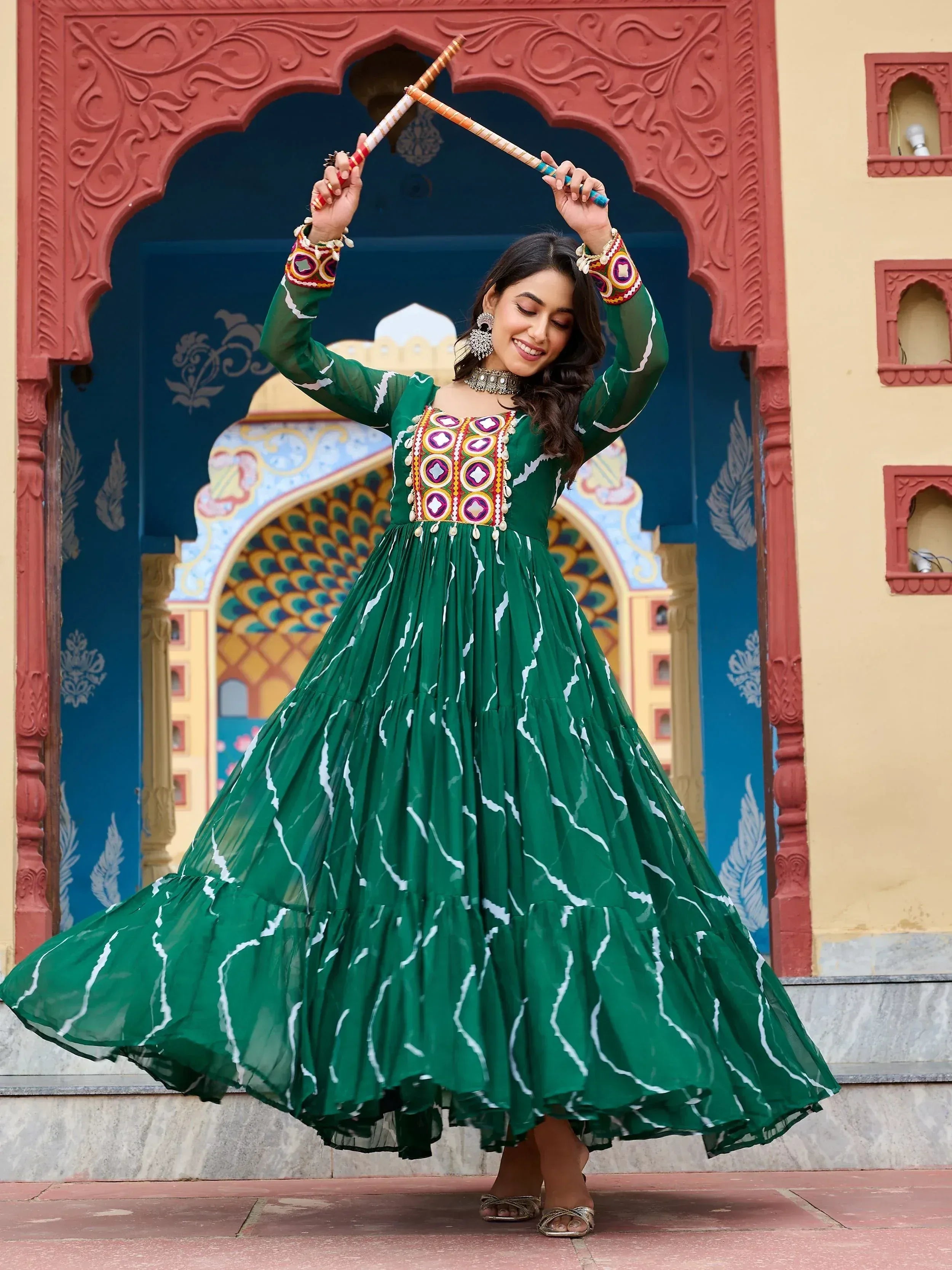 Woman in green ethnic gown with mirror work dancing with dandiya sticks against ornate Indian archway