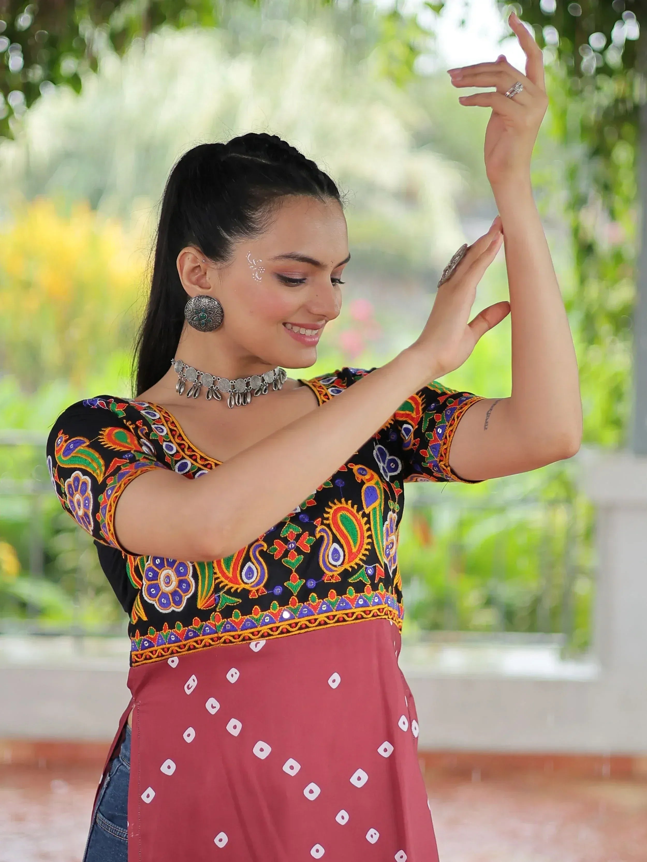 Woman in colorful embroidered Indian top with paisley design, paired with red bandhani skirt, dancing outdoors