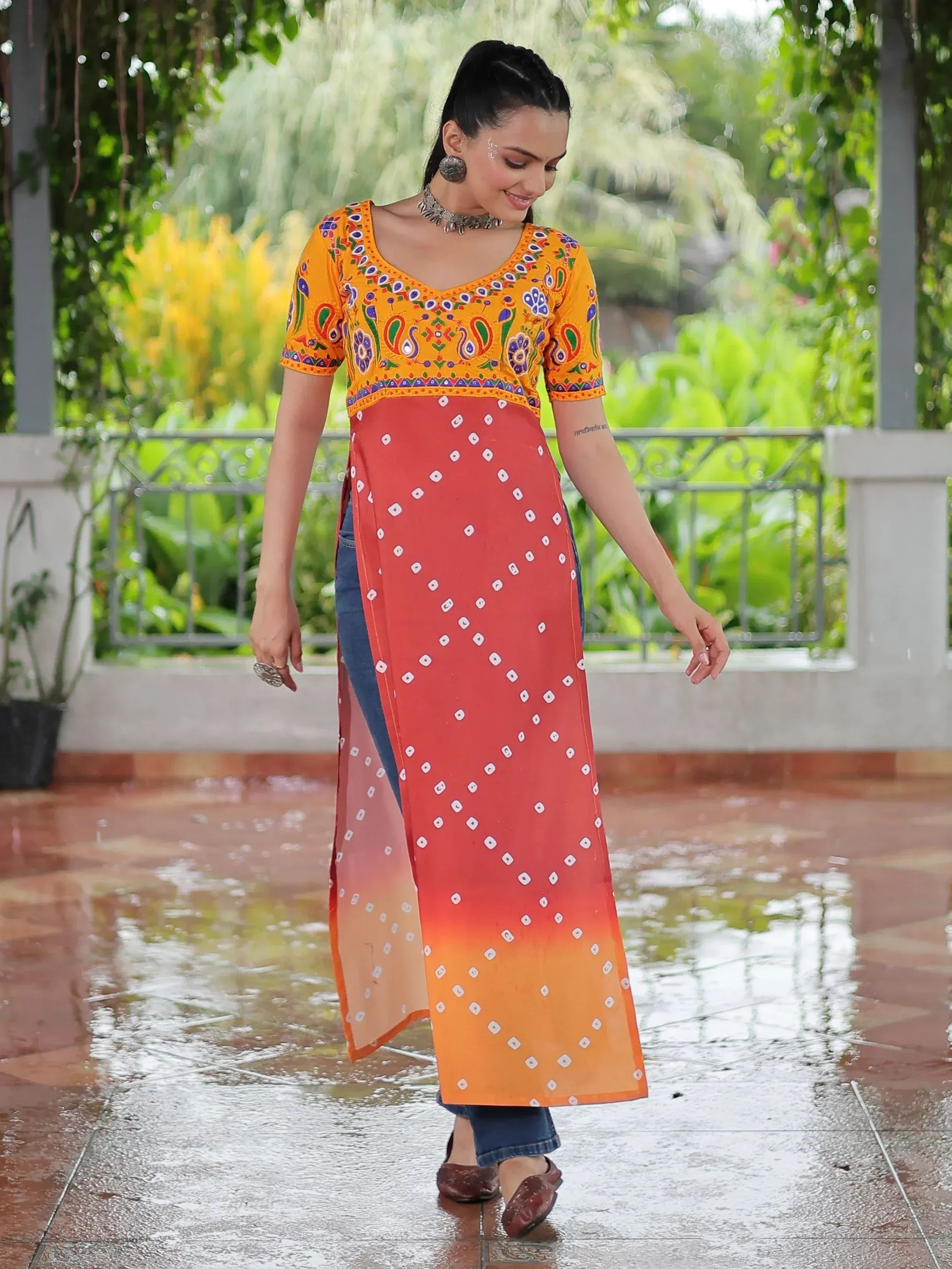 Woman in vibrant orange and red printed ethnic kurti with bandhani pattern, paired with jeans, walking outdoors