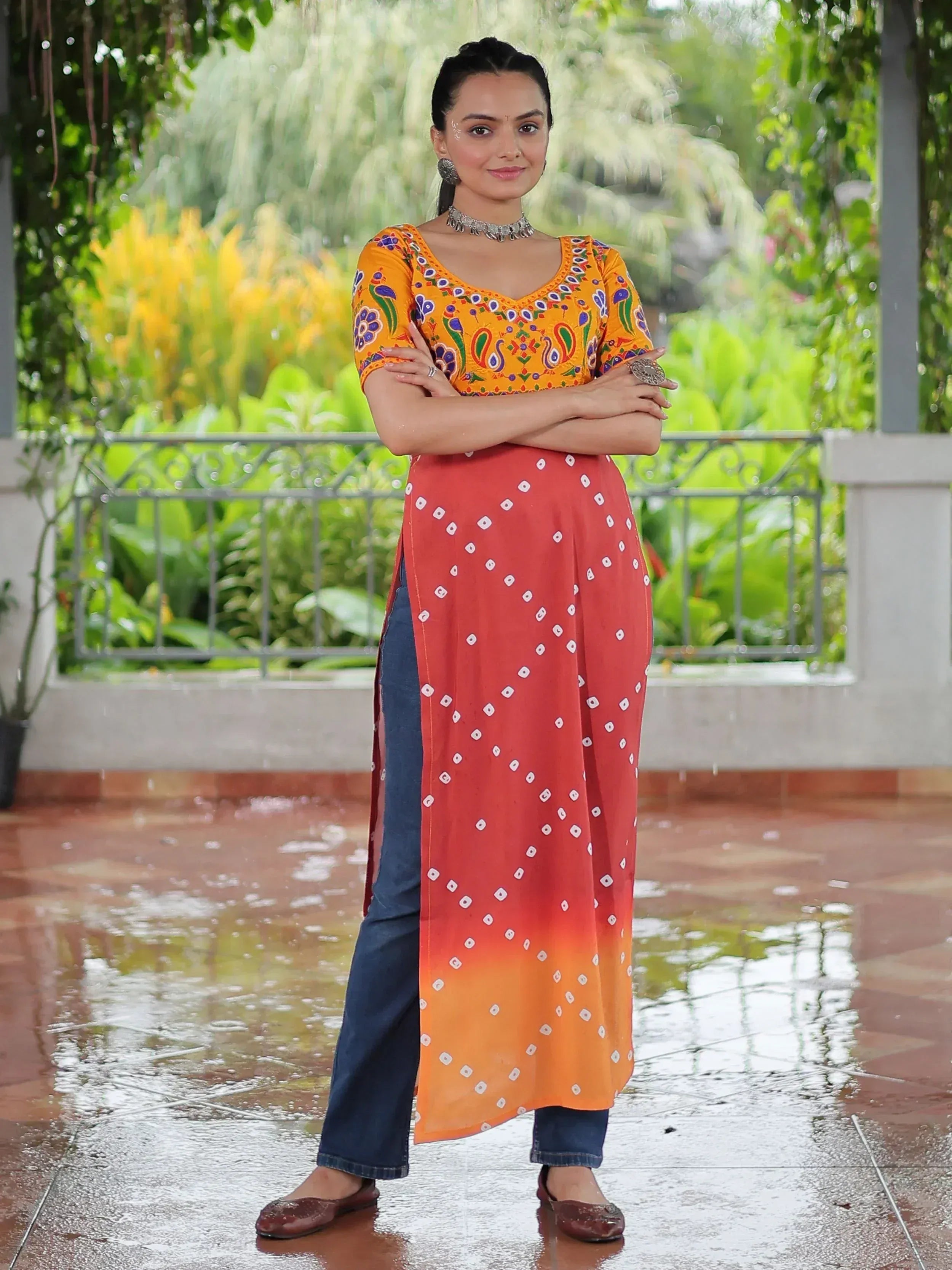 Woman in vibrant orange embroidered kurta with red bandhani drape, paired with jeans, standing outdoors in rain