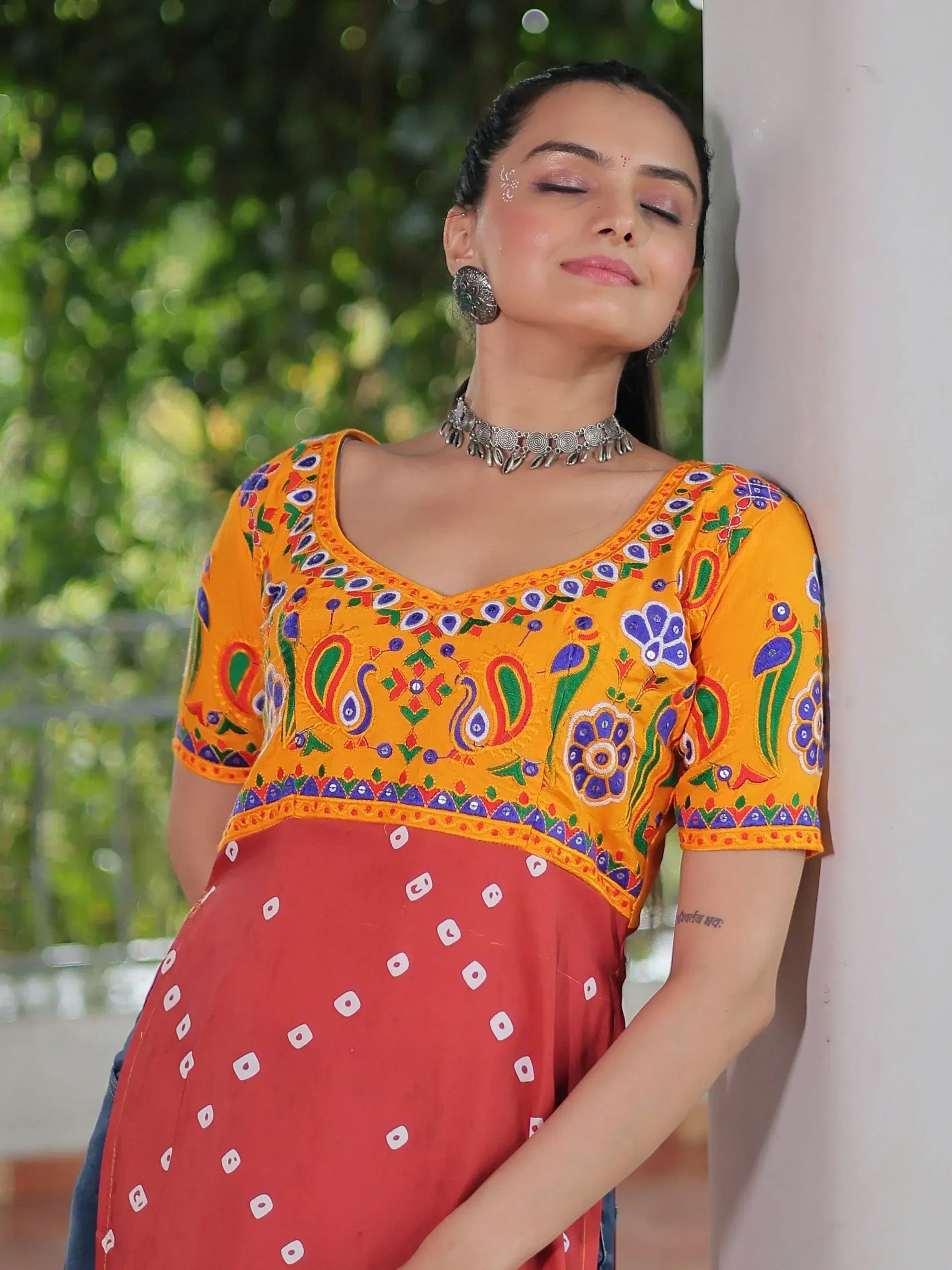 Woman in vibrant yellow embroidered top with peacock motifs and red bandhani skirt, styled with silver jewelry for ethnic wear.