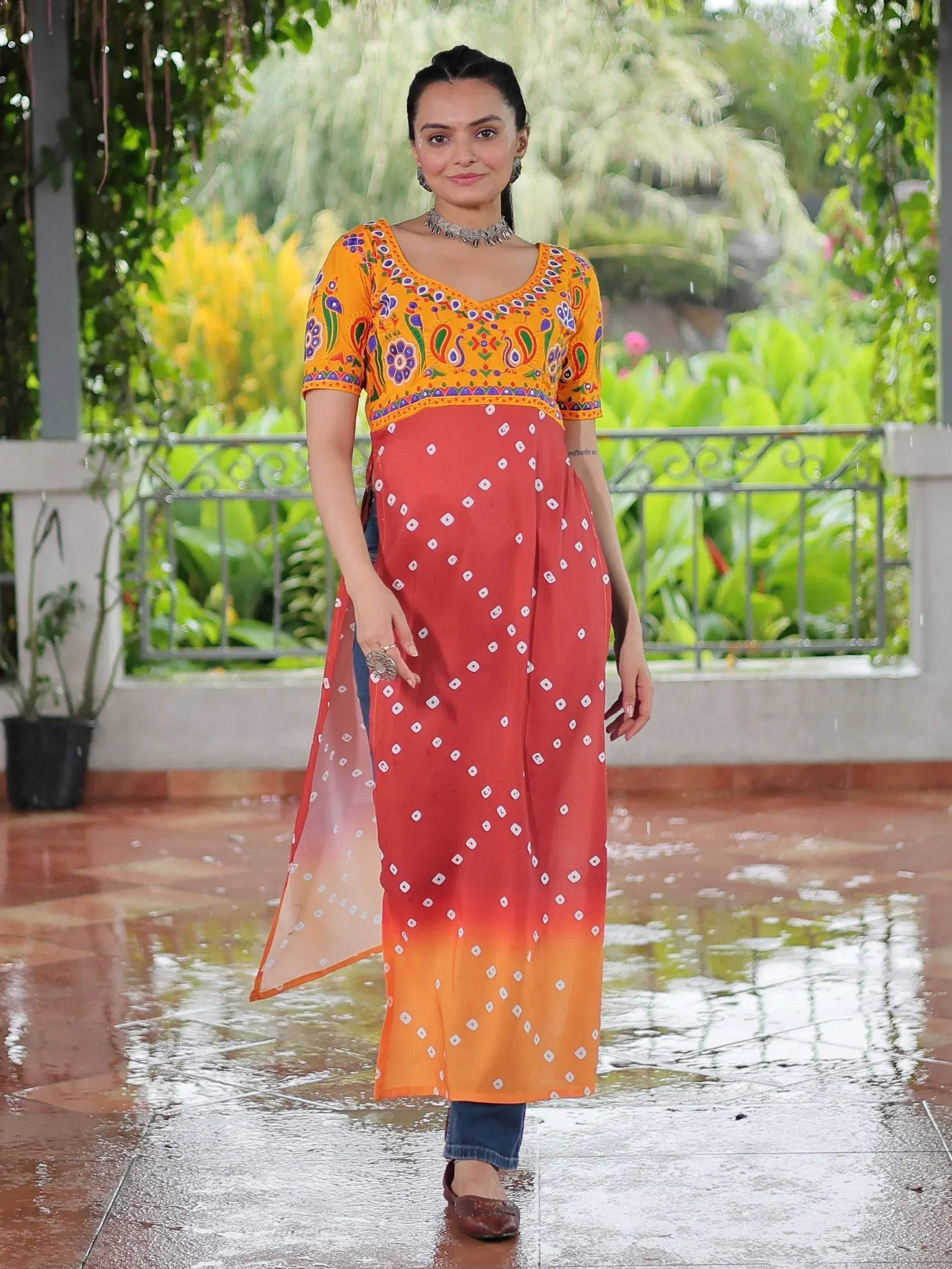 Woman in vibrant orange and red ethnic kurta with floral embroidery, paired with jeans, standing outdoors in rain.