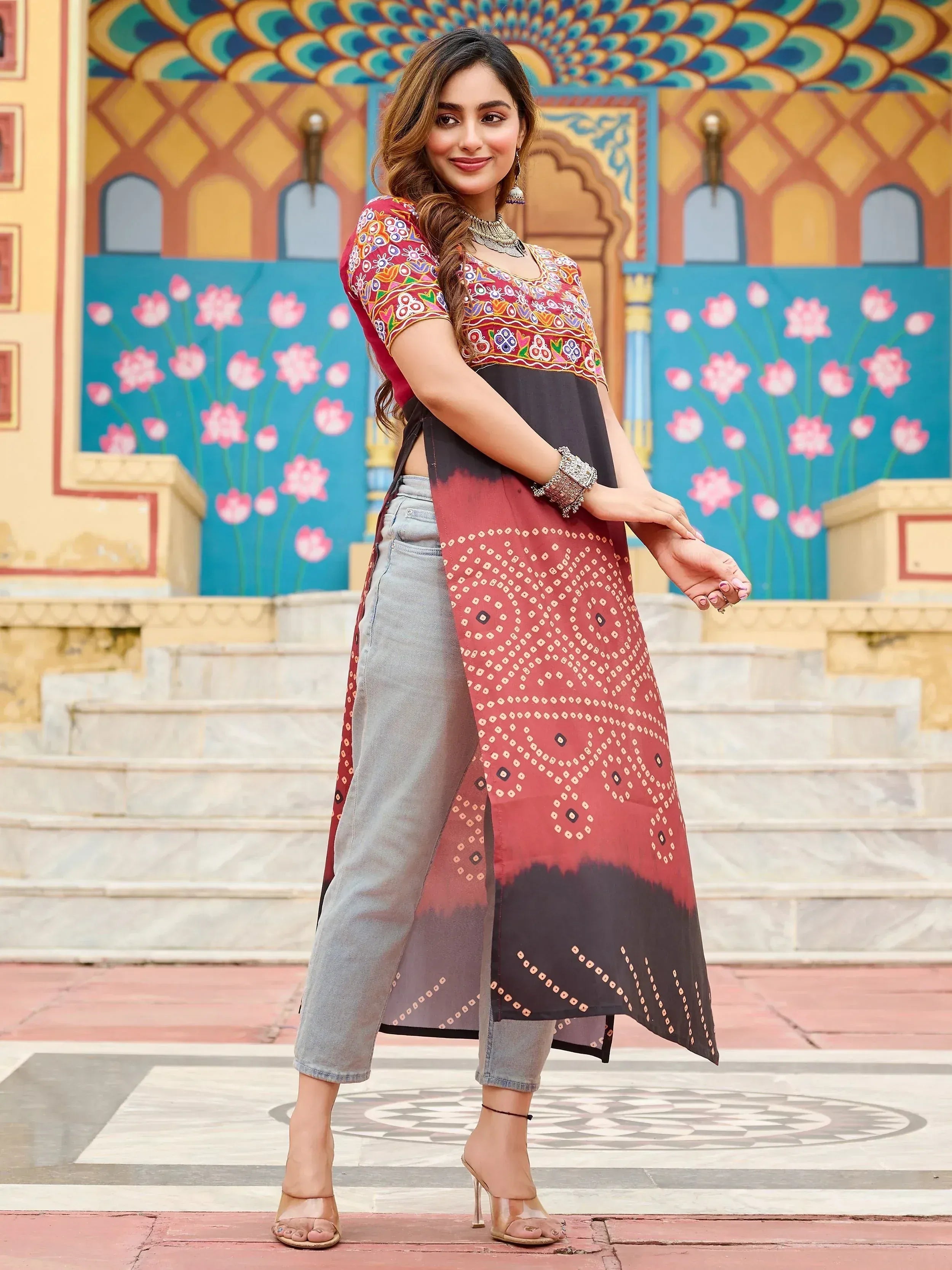 Woman in embroidered ethnic kurta with bandhani print, paired with denim pants, against colorful Indian architecture