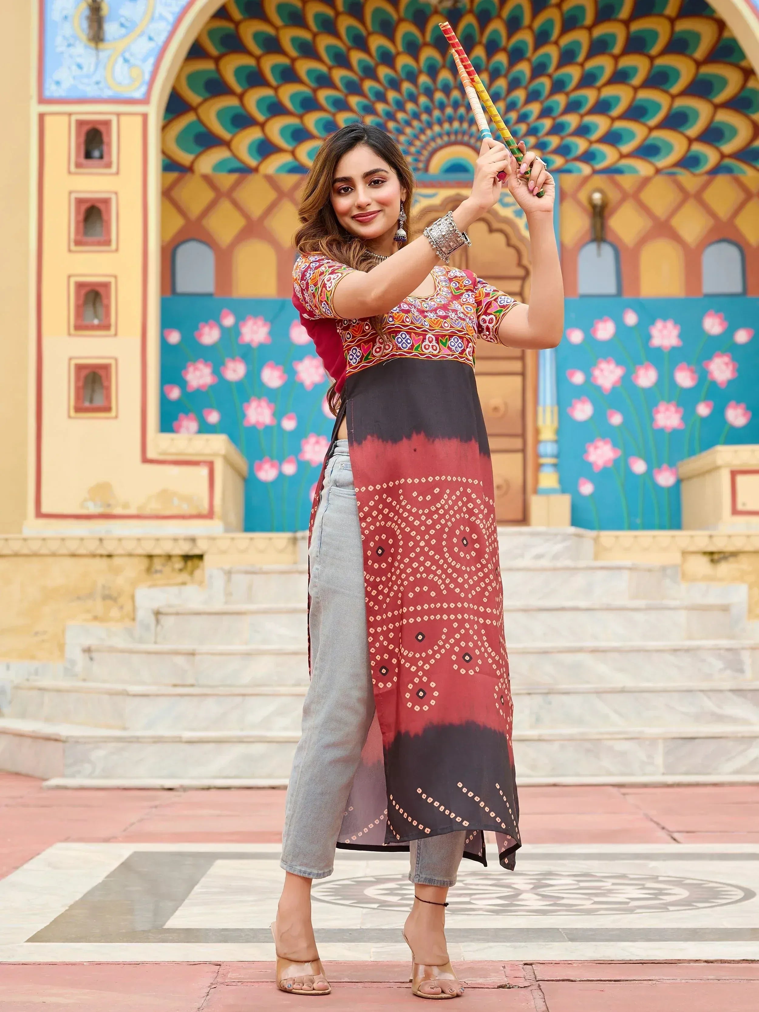 Woman in embroidered ethnic kurta with tie-dye panel, paired with jeans, holding colorful sticks against peacock mural backdrop