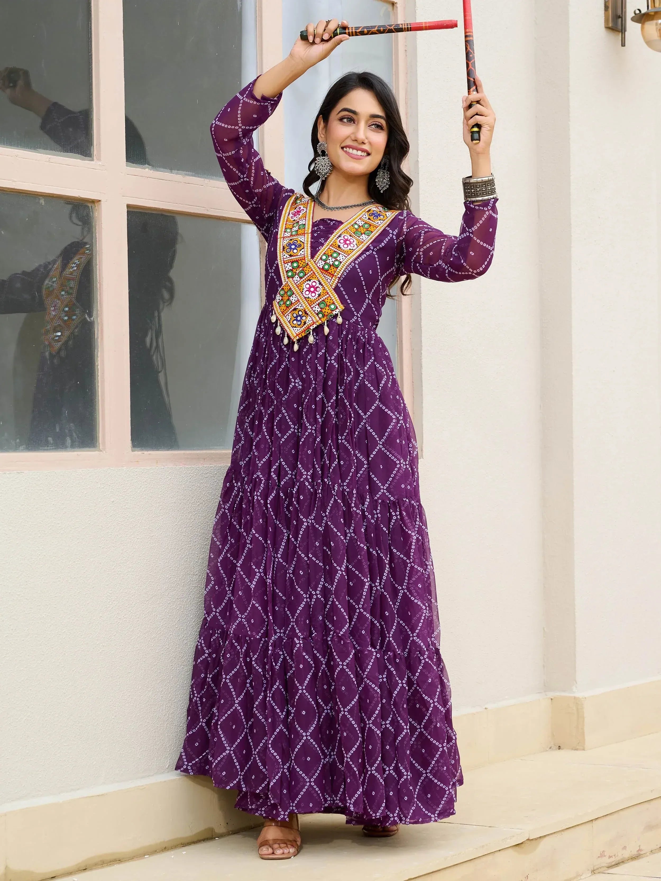 Woman in purple ethnic gown with mirror work, holding dandiya sticks, festive Indian wear